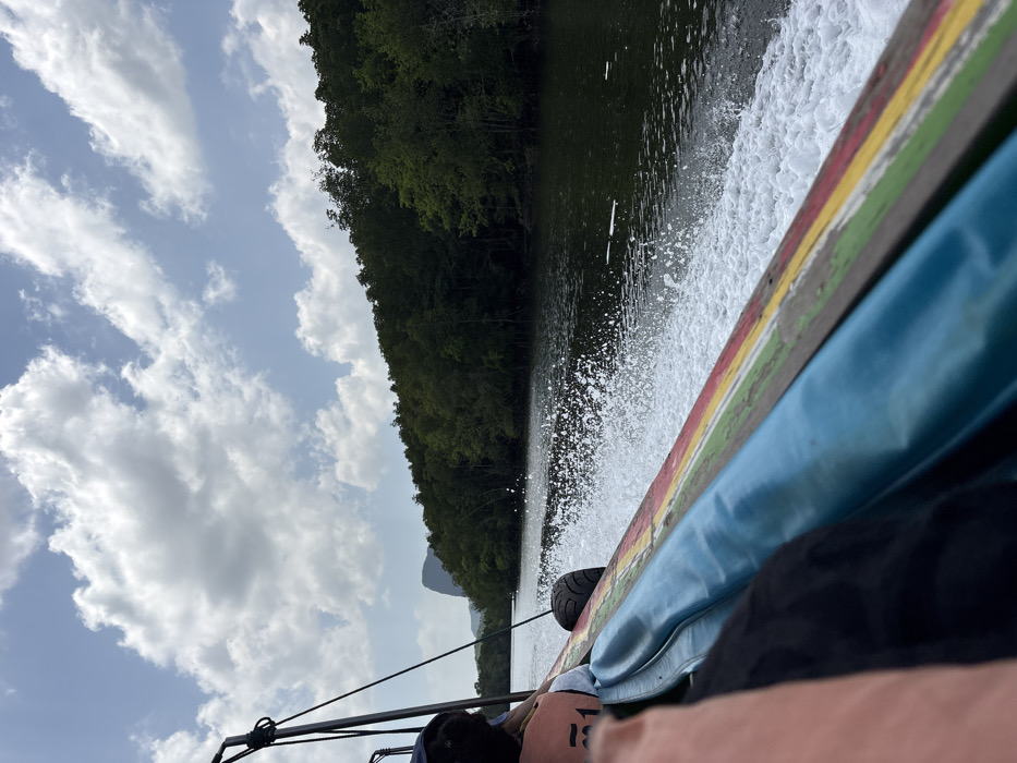 An image taken from a moving boat depicts a waterway scene in Takua Thung, Thailand. The view is angled towards the starboard side, capturing the boat's wake and the surrounding environment under a partly cloudy sky.
In the foreground, the side of a traditional Thai longtail boat is prominent, featuring horizontal stripe detailing in blue, green, yellow, and red along its wooden gunwale. A light blue padded cushion runs along the top edge of the boat's interior. A white wake of disturbed water and foam trails behind the moving vessel, indicating its forward momentum. A black tire fender is affixed to the boat's exterior. Partially visible in the bottom left are the arm and back of a passenger, with another passenger wearing an orange life vest, partially obscuring the number "151," and a dark head covering.
The waterway, with dark, calm water ahead, is flanked on both sides by dense, dark green trees, characteristic of mangrove forests. In the distant left background, a distinct, sharply peaked limestone karstic mountain is visible above the tree line. The sky overhead is predominantly light blue with numerous scattered white cumulus clouds.