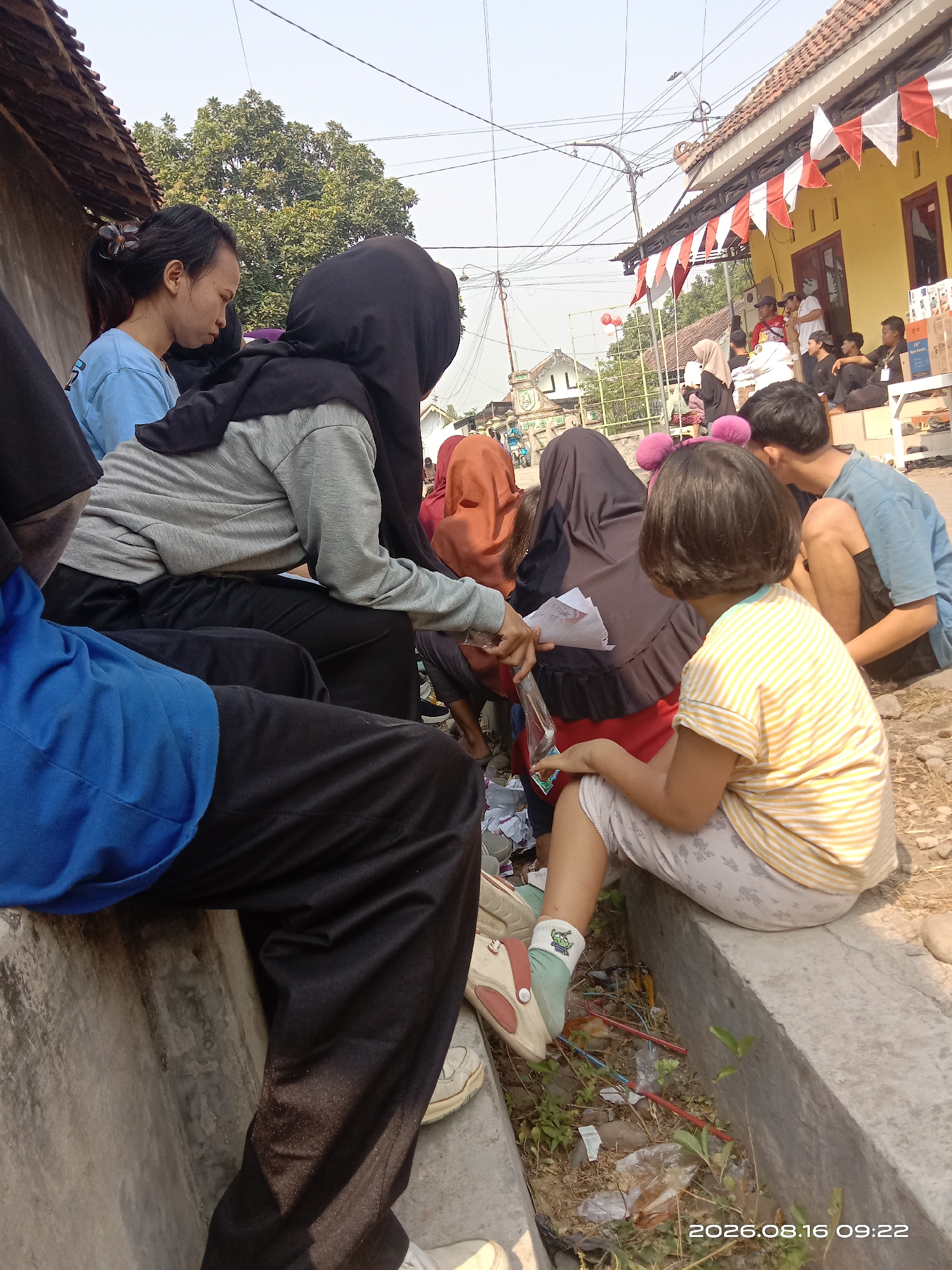 The image depicts an outdoor scene in Banyuarang, Indonesia, likely a residential area or community space. Several individuals are visible, primarily seated along a concrete drainage channel. In the foreground, a person wearing a blue shirt and black pants is partially visible, seated on the concrete. To their left, a young woman with dark hair pulled back and a flower in her hair is wearing a light blue t-shirt and looking down. Next to her, a person in a grey hoodie and black hijab is seated. Further to the right, a child with short dark hair and a yellow and white striped t-shirt and patterned pants is also seated. Behind these individuals, other people are present, some appearing to be in conversation or observing an event. Red and white pennant flags are strung across the top of a yellow building in the background, suggesting a local celebration or event. The environment appears somewhat unkempt, with debris and dried leaves visible in the drainage channel. The sky is clear and bright, indicating daylight. The timestamp "2026.08.16 09:22" is present in the lower right corner, suggesting this image was captured on August 16, 2026, at 9:22 AM.