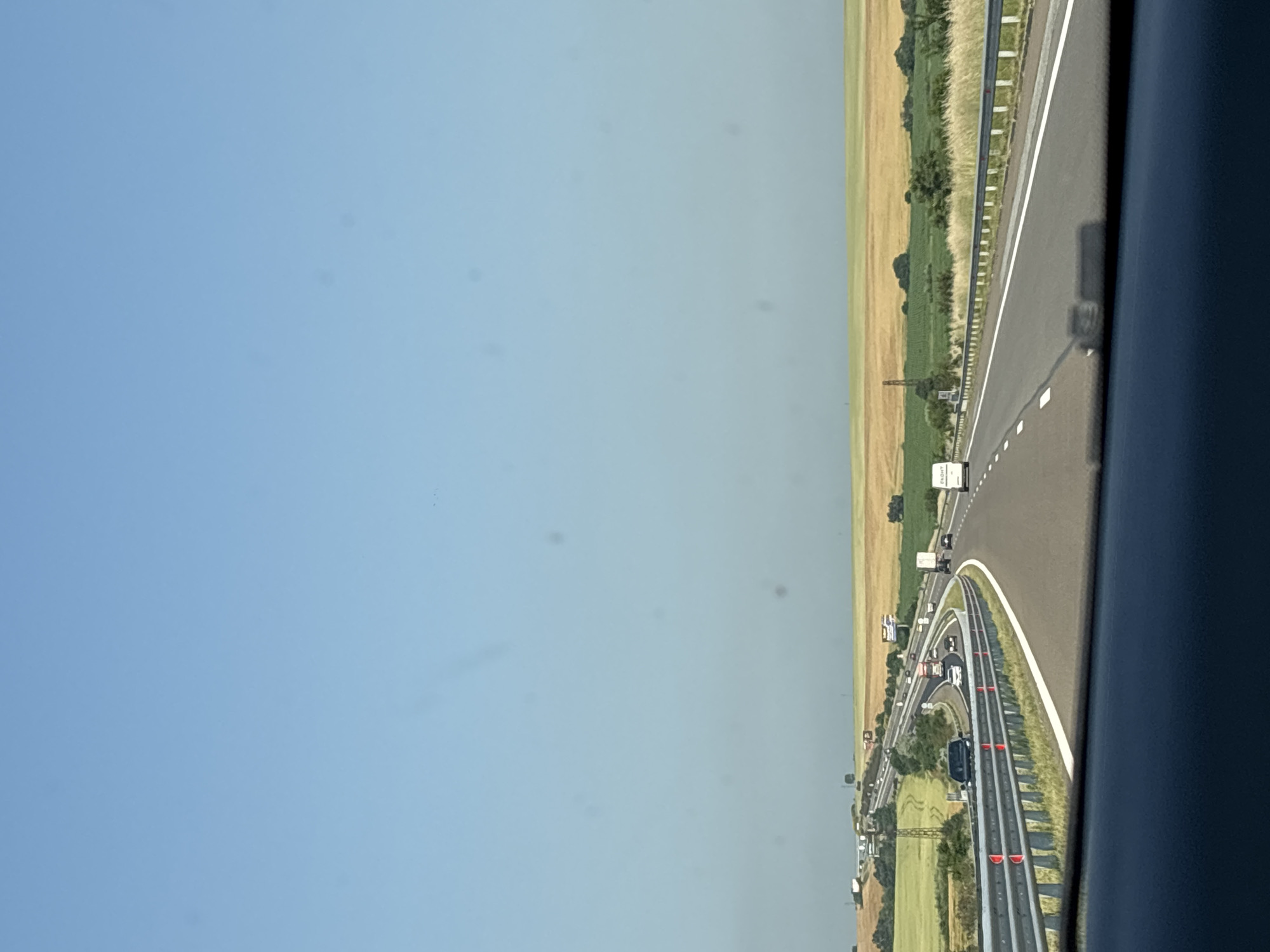 The image displays a multi-lane highway under a clear blue sky, observed from the interior of a vehicle, indicated by a dark dashboard occupying the bottom portion of the frame. Circular dust spots are visible across the sky area.
The highway, appearing to be an expressway or motorway, curves gently to the right in the foreground and features multiple lanes with distinct white markings. Metal guardrails, equipped with red reflectors, line the immediate edges of the road. Moderate vehicle traffic is present: a dark-colored SUV occupies an inner lane in the foreground, while on a higher, parallel section of the highway further to the left, multiple additional vehicles, including a red-cab truck and a white box truck, are visible. A white courier van displaying "ЕКОНТ" (EKONT) branding on its rear is positioned in the right-most lane in the mid-ground.
The surrounding landscape consists of extensive agricultural fields. Fields to the left of the highway are predominantly green, while those to the right exhibit a golden-yellow coloration. Sparse trees and bushes are scattered across the terrain. A metal lattice transmission tower is prominent in the green fields on the left side of the highway. Two billboards are also visible adjacent to the elevated road section on the left. The scene is brightly lit by daylight, indicating clear weather conditions in the Hadzhiite area of Bulgaria.