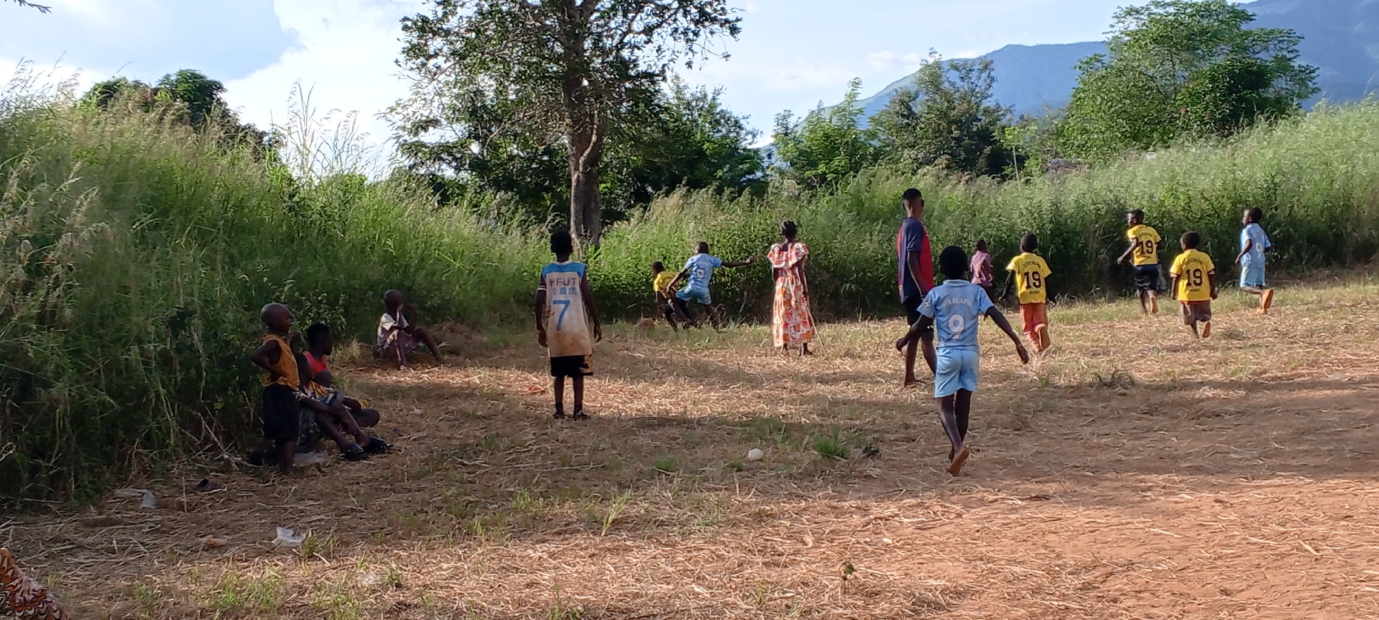 The image depicts a scene in Ngalula, Democratic Republic of the Congo, where a group of children are gathered on a patch of dry, grassy land bordered by dense foliage. Several children are seated or standing near the edge of the tall grass on the left side of the image, while others are dispersed across the open area. Some are dressed in jerseys, including yellow ones with the number 19 and blue ones with other numbers, indicating they may be engaged in a game or practice. The children's clothing and the surrounding environment suggest a rural setting. In the background, there are trees and a mountain visible in the distance.