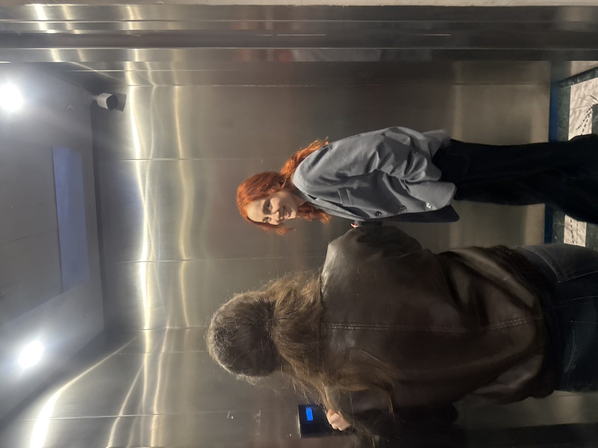 The photo shows two women in what appears to be an elevator with a stainless steel interior. The woman on the left has red hair and wears a grey blazer and black pants. She looks toward the camera with a slight smile. The other woman, closer to the elevator buttons, has long brown hair and wears a brown leather jacket. A surveillance camera is mounted in the upper left corner of the elevator. The elevator seems to be in Tbilisi, Georgia.