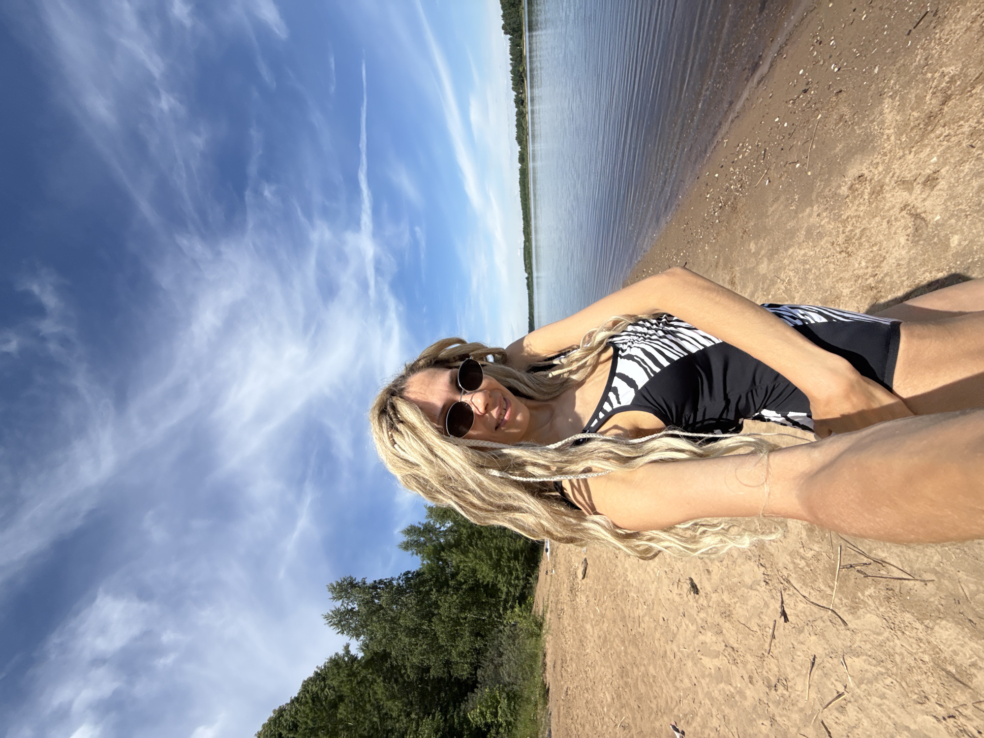 The image shows a woman with blonde dreadlocks and sunglasses standing on a sandy beach in Rusanovo, Russia. She is wearing a black and white zebra-print swimsuit. The background features a calm river with trees and a bright blue sky with scattered clouds. The woman is holding the camera, taking a selfie. The sand is light brown and there are small pieces of driftwood and shells scattered on it. The water is dark blue and reflects the sky. The trees in the background are dense and green, suggesting a wooded area. The overall scene depicts a sunny day by the river.