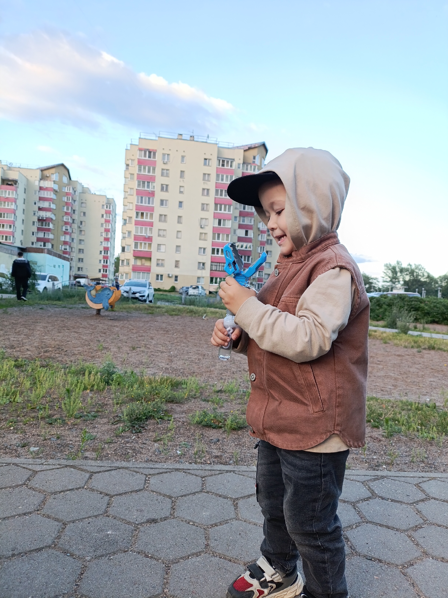 A young boy, wearing a brown hooded vest over a tan long-sleeved shirt, dark jeans, and sneakers, stands on a hexagonal paver pathway. He is looking down and smiling, holding a blue bubble gun in his right hand and a small bottle of bubble solution in his left.
The background features apartment buildings with pink and white facades under a partly cloudy sky. The time of day appears to be late afternoon or early evening, given the soft lighting. The setting is an outdoor area with patches of grass and dirt, suggesting a park or a residential courtyard. In the mid-ground, there is a yellow and orange playground ride shaped like a whale, and several parked cars. A person in a black jacket and pants is walking away from the camera on a paved path in the far left background. The overall mood is cheerful and playful. There is no visible text in the image.