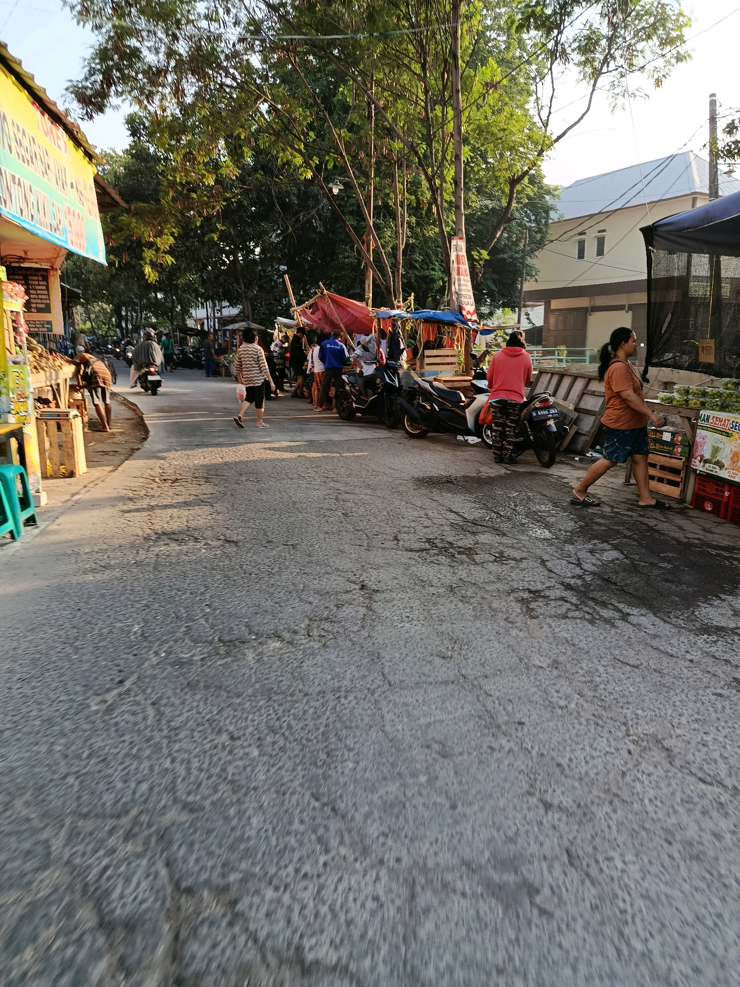 The image depicts a street scene in Bekasi, Indonesia, likely a market area, characterized by stalls and pedestrian activity. In the foreground, the asphalt road shows signs of wear with cracks and patches of moisture. To the left, a brightly colored yellow stall with text in Indonesian is visible, alongside wooden crates. Further down the street, several market stalls with red and blue tarps are set up, displaying goods.
Various individuals are present, engaged in different activities. On the left side, a person is seen near a stall with what appear to be fruits or vegetables. Further back, people are walking and interacting near the stalls. In the center of the frame, a person in a striped shirt walks away from the camera, carrying a plastic bag. To the right, a person in a pink hoodie and patterned pants stands next to a motorcycle. Another woman in an orange shirt and patterned skirt walks towards the right of the frame, past a stall with crates of green produce and promotional posters. Several motorcycles are parked or being ridden on the street, indicating local transportation.
The background features trees with lush green foliage casting shadows on the road. Beyond the trees, a light-colored building with a sloped roof and windows is visible, suggesting it is a residential or commercial structure. The overall atmosphere appears to be that of a bustling daytime market with moderate pedestrian and vehicle traffic under a clear sky.