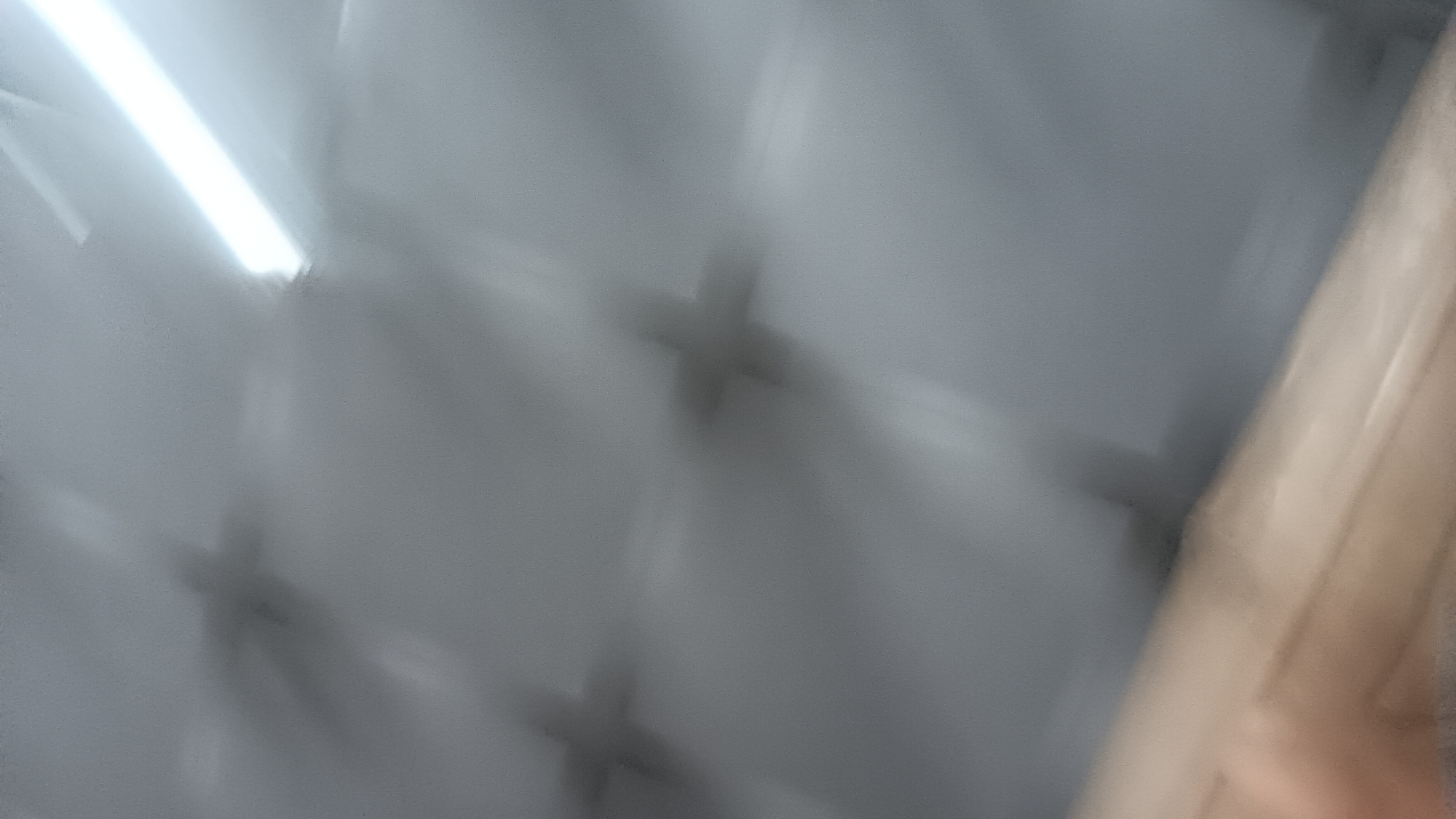 The image depicts a blurry, low-light interior scene, possibly a ceiling with reflective surfaces and shadows. Several dark, cross-shaped shadows are visible, suggesting objects casting these shapes. In the lower portion of the image, a blurred, light-colored surface is present, appearing to be made of wood or a similar material. The overall impression is that of an industrial or storage space, with the cross shapes possibly being parts of overhead lighting fixtures or structural elements. The location in Sonobekel, Indonesia, is not discernible from the visual information provided.