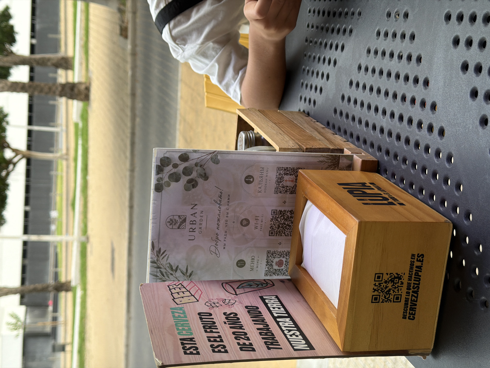The image shows a close-up of a table at an outdoor establishment in Urbanització Cales I Talaies, Spain. In the foreground, a wooden napkin dispenser holds white napkins. The dispenser is branded with "LUPIA" and a QR code that links to "CERVEZASLUPIA.ES." To the left of the dispenser is a sign with Spanish text and a graphic of a beer glass. To the right of the dispenser, a menu from "URBAN GARDEN" is displayed, featuring text in Russian and QR codes for menu access, Wi-Fi, and a hookah menu. The menu also includes a welcome message in Russian. A person, partially visible in the upper right, is seated at the table, wearing a white shirt and a black strap. The table itself is dark gray with a perforated surface. In the background, trees and a building are visible, suggesting an outdoor seating area.