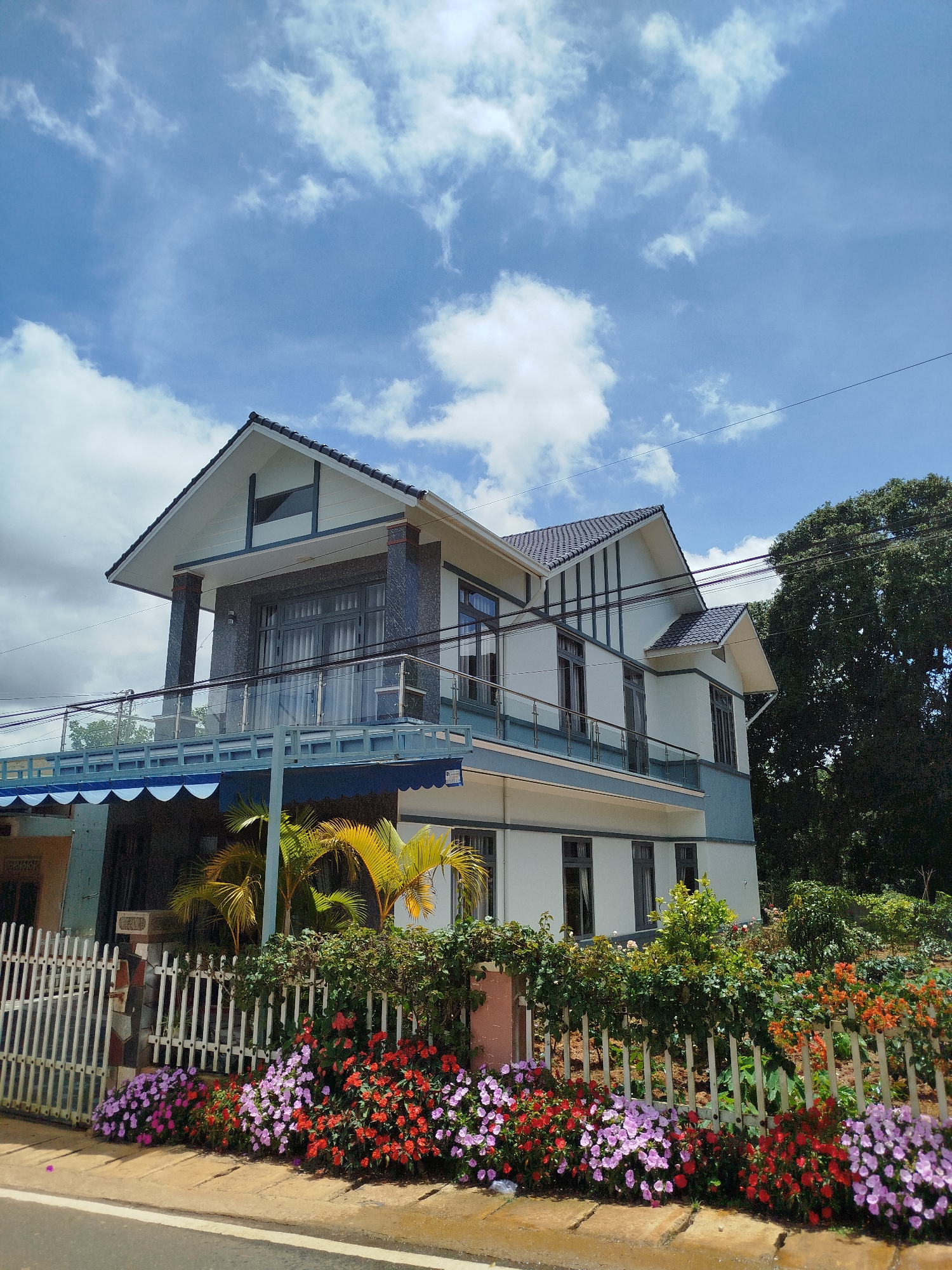 The image depicts a modern, multi-story house situated in Trường An, Vietnam. The house has a white and blue exterior with dark-tiled roofing. It features a prominent balcony with glass railings, and a blue awning extends from the front of the house. In front of the house, a white picket fence encloses a vibrant garden filled with blooming flowers in shades of pink, red, and orange. Several palm trees are also visible within the property. The environment suggests a bright, sunny day with a partly cloudy sky. Power lines run across the top of the image, and a paved road is visible in the foreground. There are no people or animals present in the image. The architectural style and the surrounding vegetation are characteristic of a residential area in Vietnam.