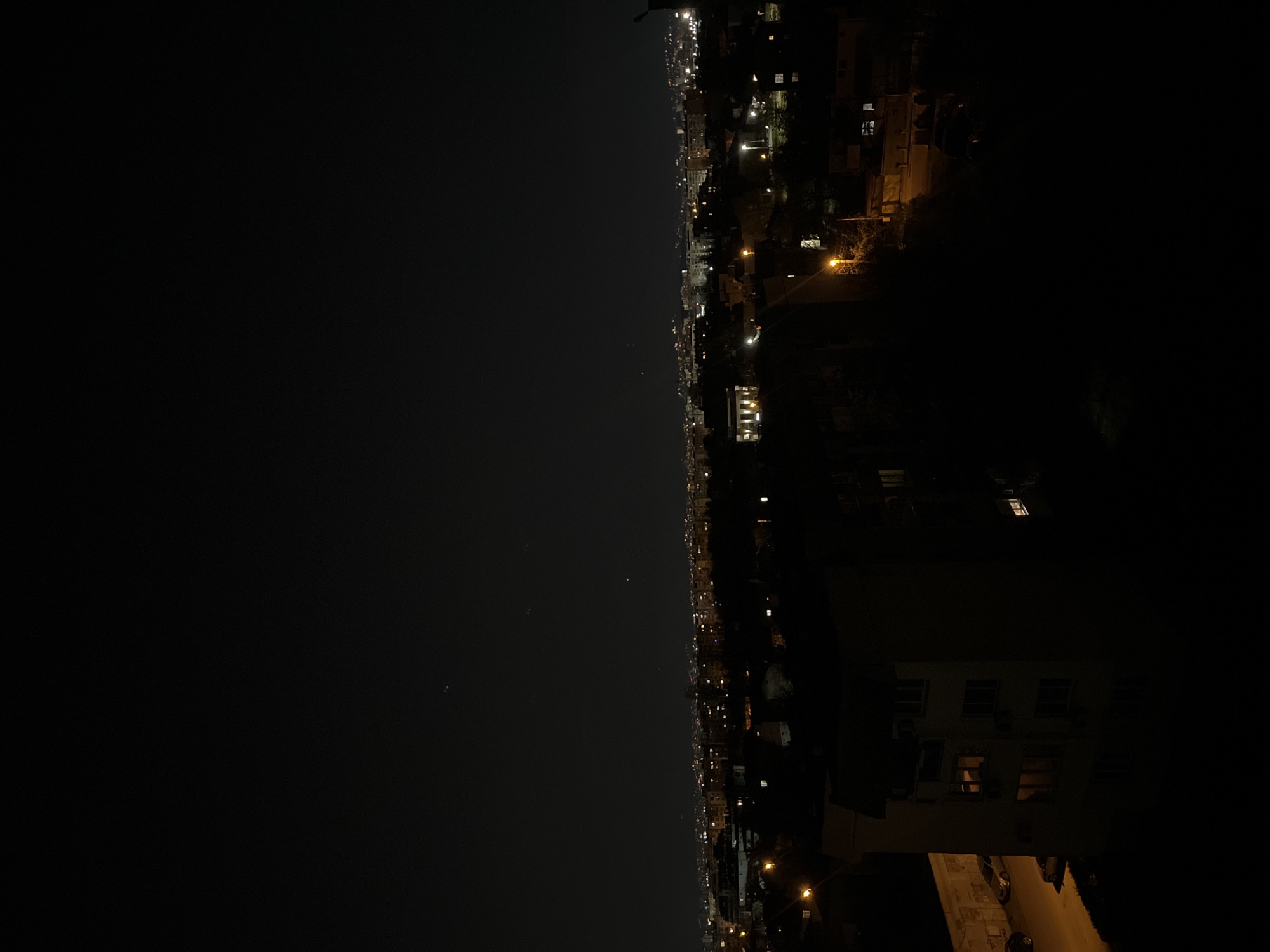 The image captures a nighttime view of Sofia, Bulgaria. The city stretches across the right side of the frame, with numerous buildings illuminated by streetlights and the lights from windows. The left side of the frame is dark, likely depicting the sky. The angle suggests the photo was taken from a high vantage point. No people or specific activities are discernible due to the distance and darkness.