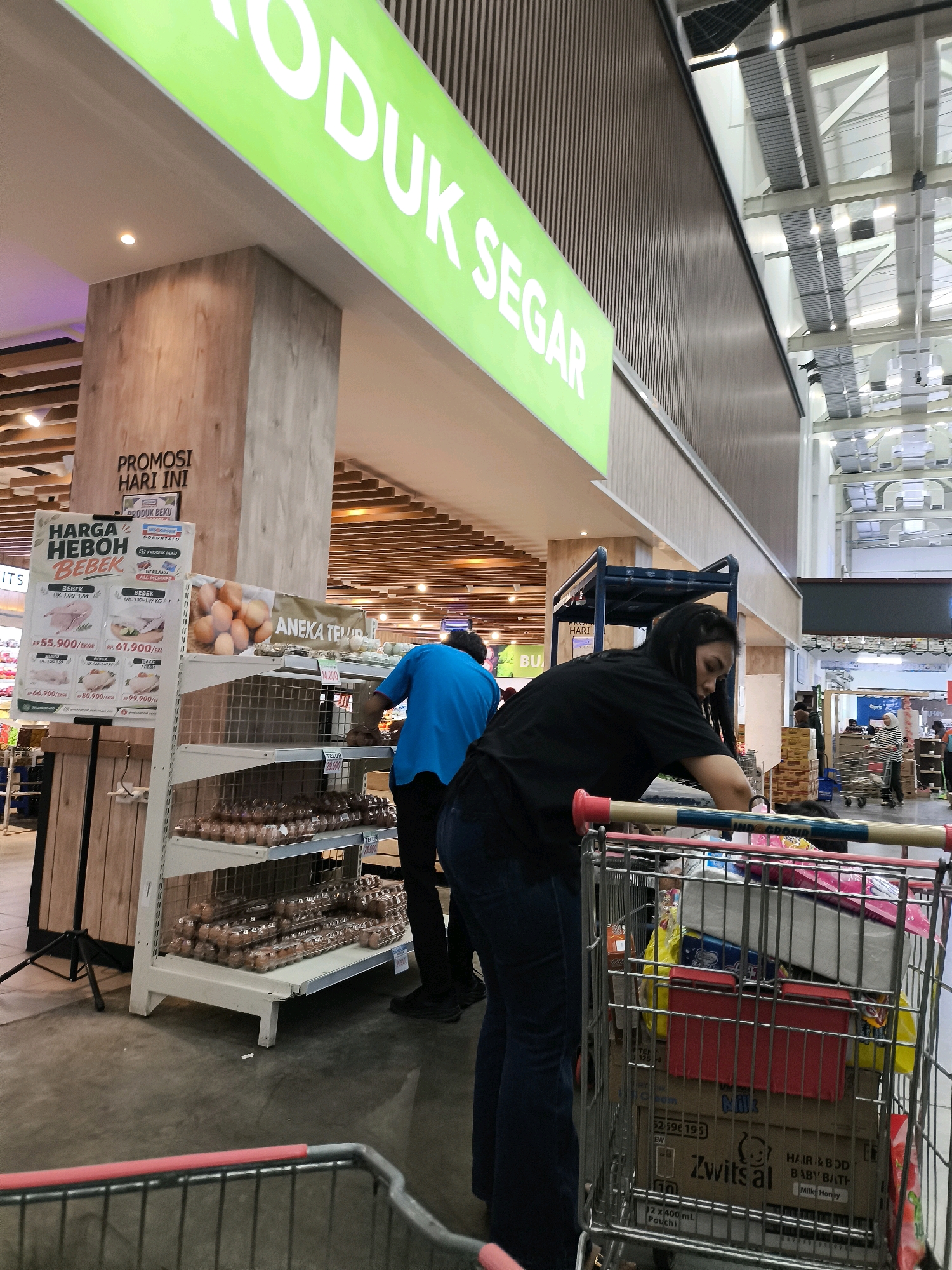 This is a medium shot, eye-level, indoor photograph taken at a supermarket in Gorontalo, Indonesia. The image features a grocery shopping cart in the foreground, filled with various products, including boxes of "Zwitsal Baby Bath Milk Honey" and other items. A woman wearing a black t-shirt and blue jeans is standing behind the cart, pushing it. Another person, wearing a blue t-shirt and black pants, is visible to the left, appearing to be organizing or stocking items on a shelf. In the background, a large green sign with white text, possibly reading "PRODUK SEGAR" (Fresh Product), is prominently displayed. Shelves stocked with numerous cartons of eggs are also visible. The supermarket environment appears modern with high ceilings and overhead lighting. The floor is a concrete surface. The overall scene depicts a typical shopping experience in a grocery store.