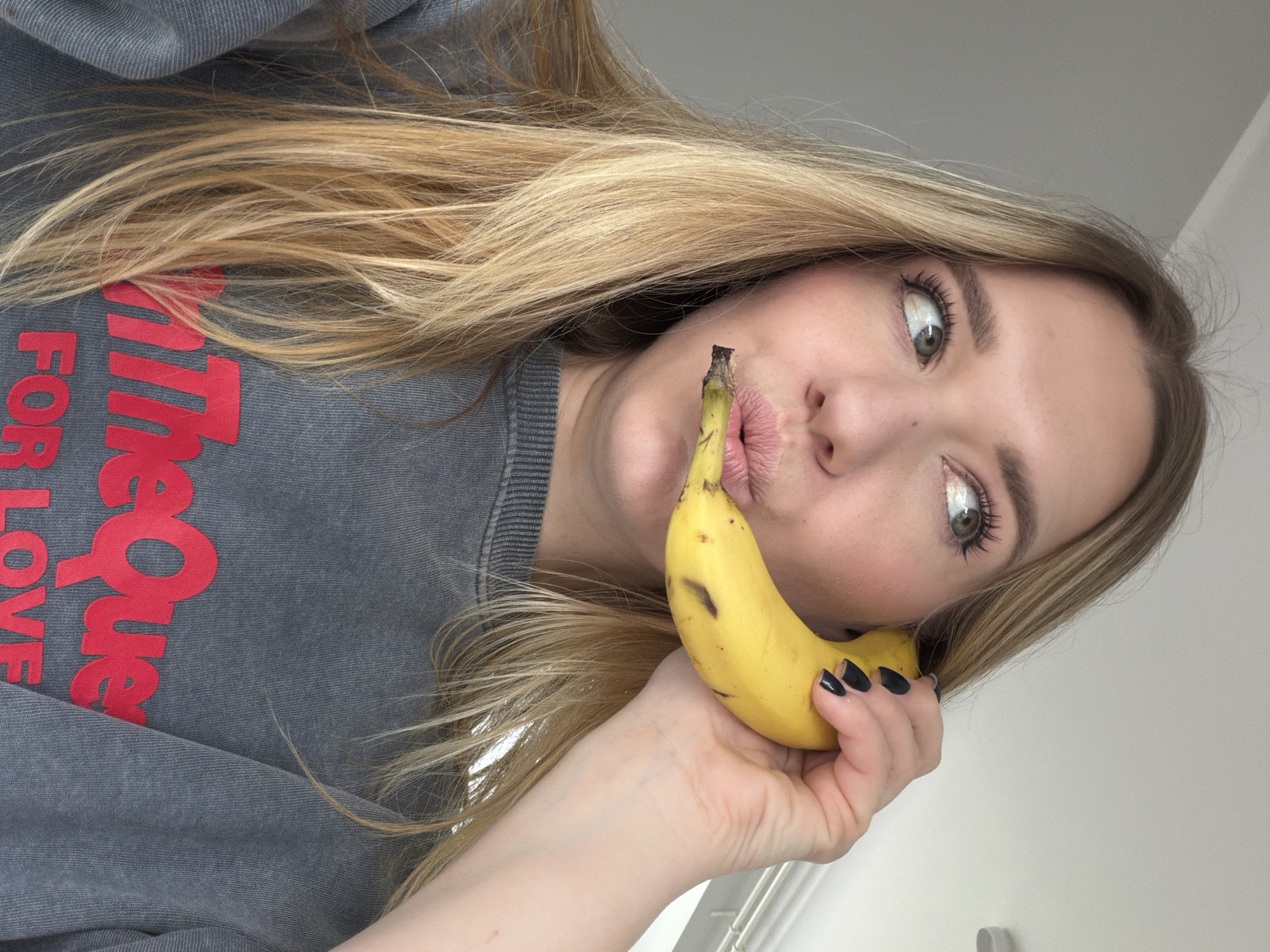 A blonde woman is holding a banana near her mouth, as if posing for a selfie. She's wearing a grey sweatshirt with red lettering that reads "The Love For" vertically, though the last word is cut off by the edge of the image. She has fair skin, green eyes, and wears makeup including mascara and blush. Her fingernails are painted black. The background is blurred and indistinct, showing a white wall and what might be a window or pipe. It is not possible to determine if the image was taken in the city of Dreiliņi, Latvia.