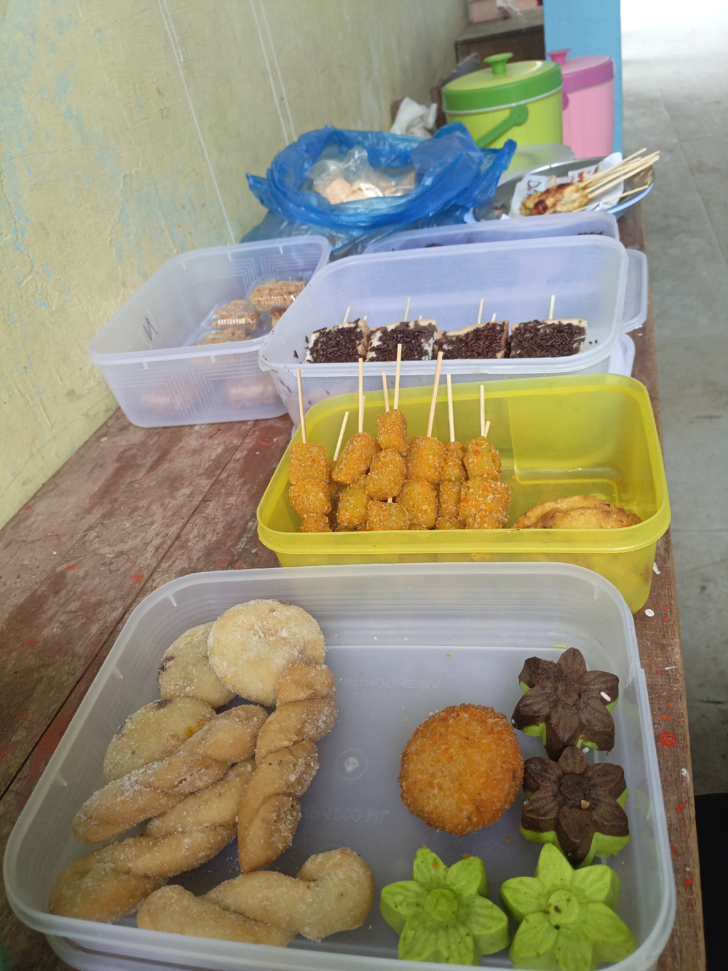 The image depicts an outdoor food stall in Meunasah Raya, Indonesia, likely on a wooden table. In the foreground, a transparent plastic container holds an assortment of snacks. These include twisted dough pastries dusted with sugar, round fried fritters, and flower-shaped cookies in chocolate, green, and a mix of colors. Behind this container, a yellow plastic container holds skewered, breaded fried food items, possibly chicken or fish, and a few pieces of what appear to be fried bananas. Further back, another transparent container displays what seems to be thinly sliced bread topped with chocolate sprinkles and skewered with toothpicks. Another similar container holds assorted cookies. To the right, a plate contains skewered food items, one of which appears to be a hot dog wrapped in pastry. In the background, two large plastic bins, one green and one pink, are visible, along with a blue plastic bag filled with food items. The overall setting appears to be casual, with a focus on various street food offerings.