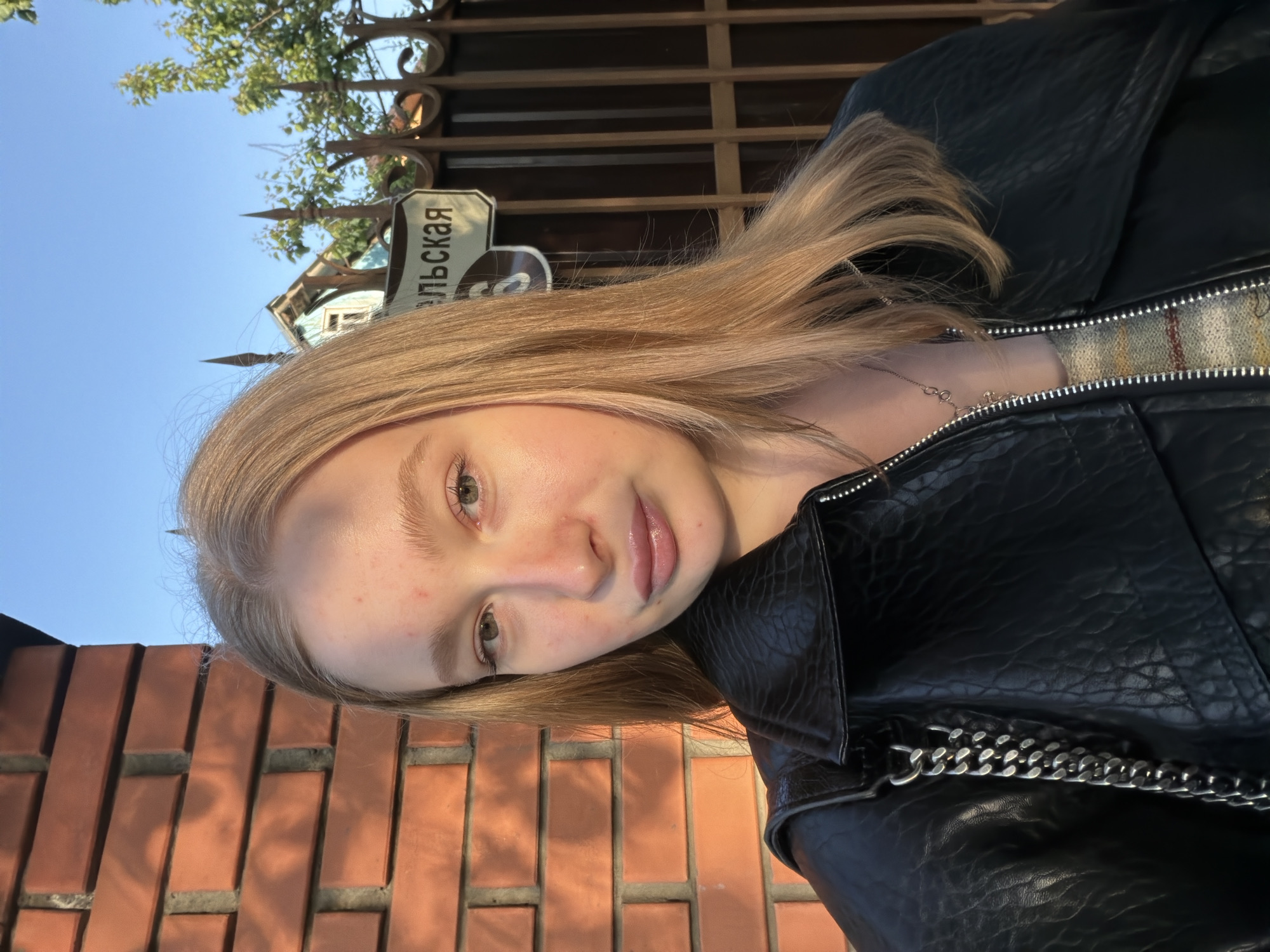The image shows a young woman with blonde hair wearing a black leather jacket. She is standing outside, with a brick wall to her left. Above her is a street sign in Russian that reads "вильская", indicating she is in the city of Beryozovsky, Russia. Behind her, there is a fence and some greenery, suggesting a residential or park area. The lighting indicates it is daytime.