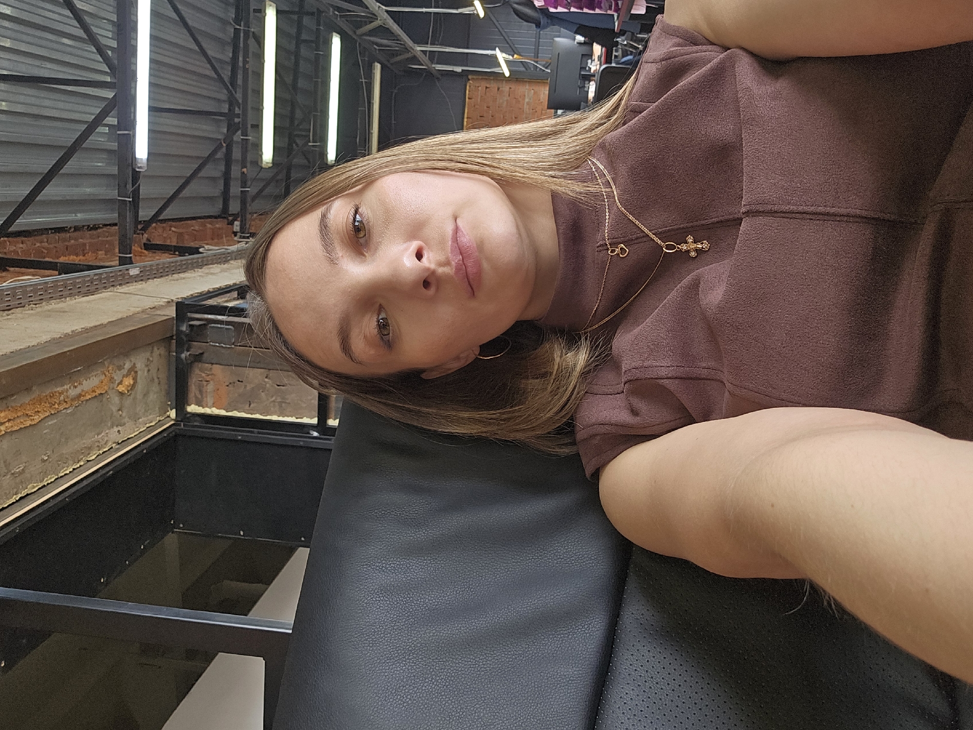 The image shows a woman with light brown hair, wearing a brown top and a gold necklace with a cross pendant. She is resting on a black leather surface, possibly a piece of furniture. The background reveals a partially constructed or industrial-looking space with metal beams, exposed brick, and artificial lighting. The location is assumed to be Chaikovsky, Russia.