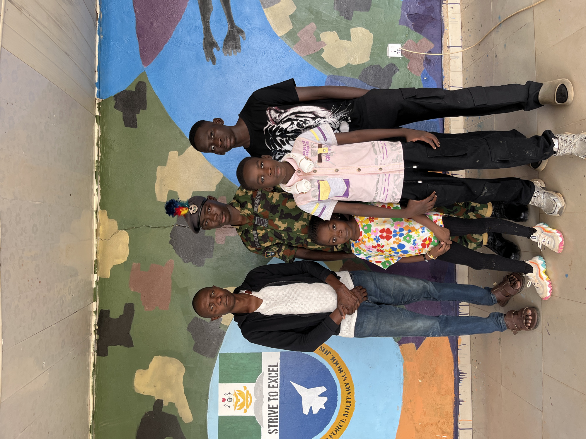 In Bar Jirgi Summa, Nigeria, in front of a mural at the Air Force Military School Jos, a group of five individuals is posing for a photograph. From left to right, a man in a black zip-up jacket over a white V-neck shirt and blue jeans stands with his hands clasped. Next to him, a Nigerian Air Force officer in camouflage uniform and a beret stands tall. In front of the officer, a young girl in a brightly colored floral t-shirt and black pants looks towards the camera. To her right, a boy in a light pink collared shirt with multicolored accents and black cargo pants, wearing headphones around his neck, also faces forward. Completing the group on the far right is another young man in a black t-shirt with a tiger graphic and black pants. The mural behind them features a camouflage pattern, a crest with an airplane and the words "STRIVE TO EXCEL" and "FORCE MILITARY SCHOOL JOS," and abstract shapes in blue, purple, and orange. A white electrical outlet with a cord is visible on the wall to the right. The floor appears to be tiled or concrete.