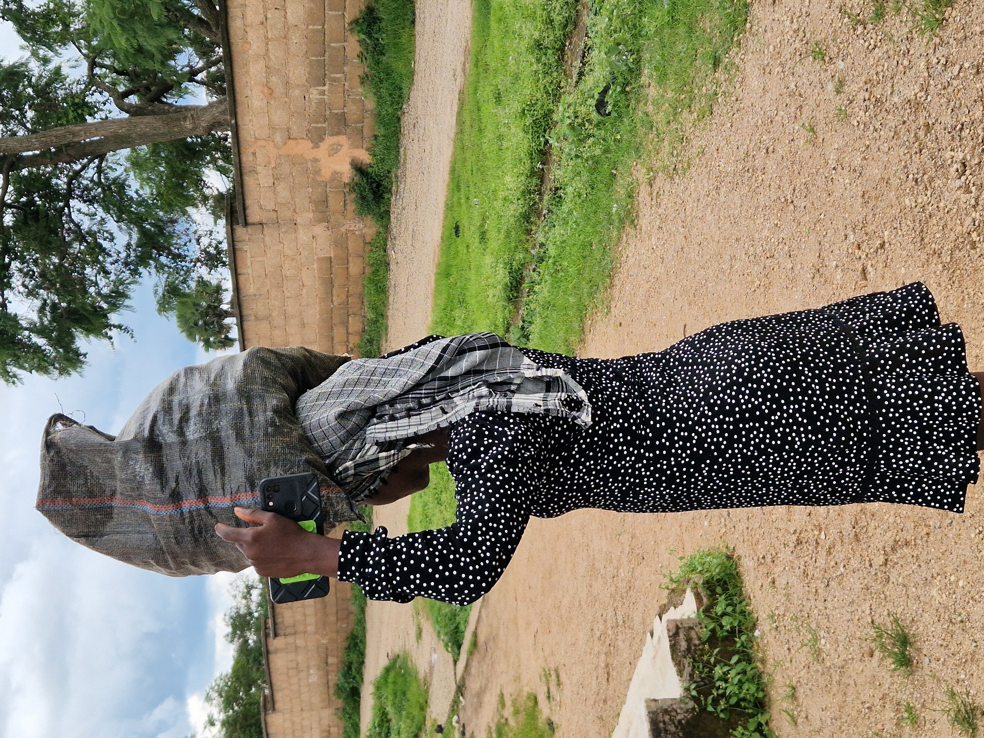 A person, viewed from the side and slightly from behind, carries a large, dark gray burlap sack on their head. The sack has red and blue stripes near its opening. The person wears a black dress with small white polka dots and a patterned headscarf. They hold a smartphone in their right hand, near the sack. The scene is outdoors, with a dirt path in the foreground and a low brick wall in the background. Lush green grass and trees are visible. The sky is partly cloudy. This appears to be a typical scene in a residential area of Jos, Nigeria, where such everyday activities are common.