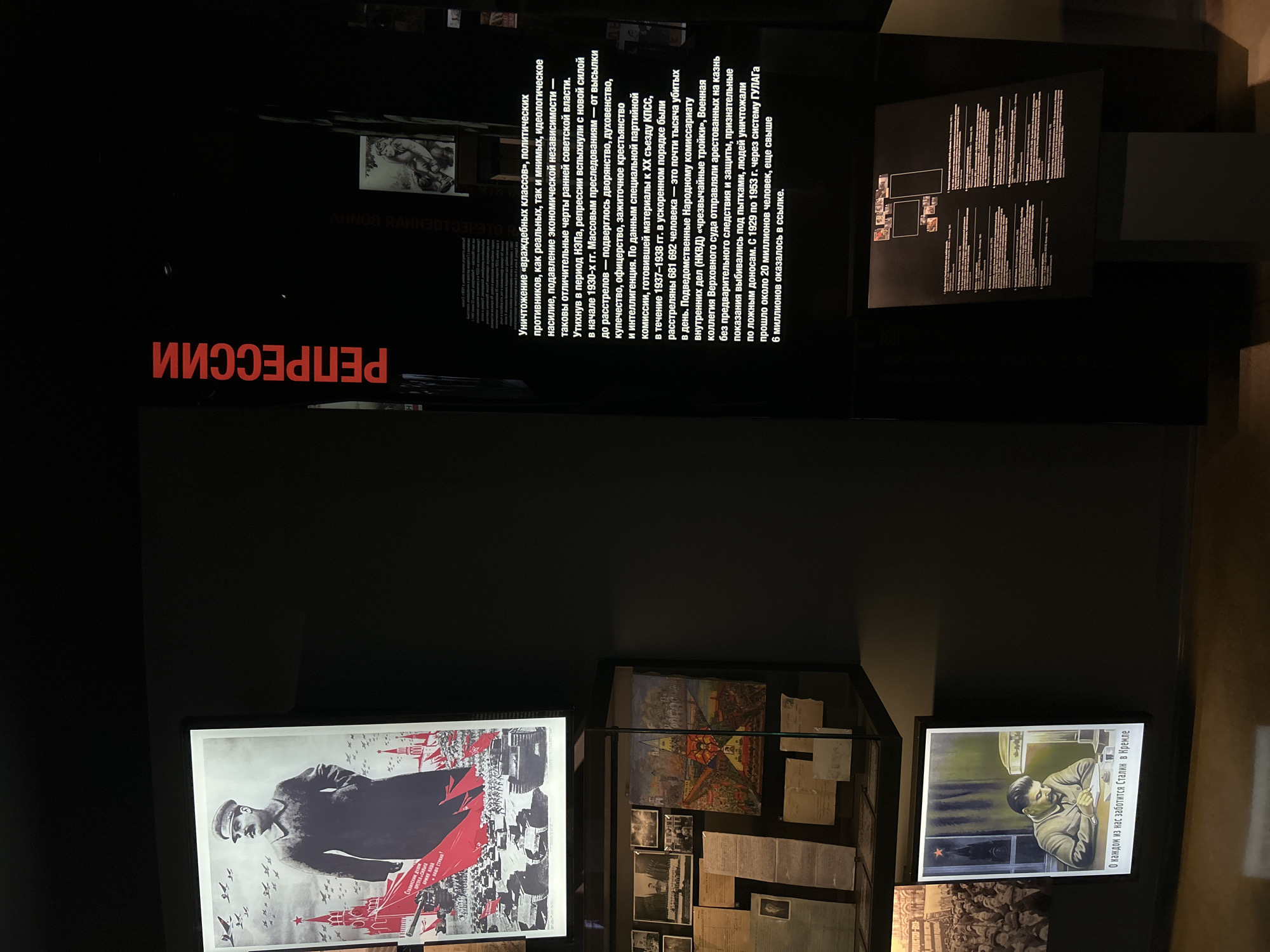 The image depicts an exhibition display about Soviet repression. The display features several elements including a large poster on the left depicting Joseph Stalin and a military parade, with red banners and tanks. Below this poster is a display case containing photographs, documents, and artwork, one of which shows a stylized depiction of a city. To the right of the main poster, vertical red text reads "РЕПРЕССИИ" (Repressions). Further to the right, there is informational text in Russian on a black background discussing the mass repressions and their victims. Below this text is another display board with smaller images and text. At the bottom of the image, to the left, is another poster with an illustration of Stalin at a desk, and text below it. The environment appears to be a museum or exhibition hall, with dark walls and spotlights illuminating the exhibits. The scene is indoors, likely in a dimly lit space to highlight the displays. There is no visible indication of the specific location within Yekaterinburg, Russia.