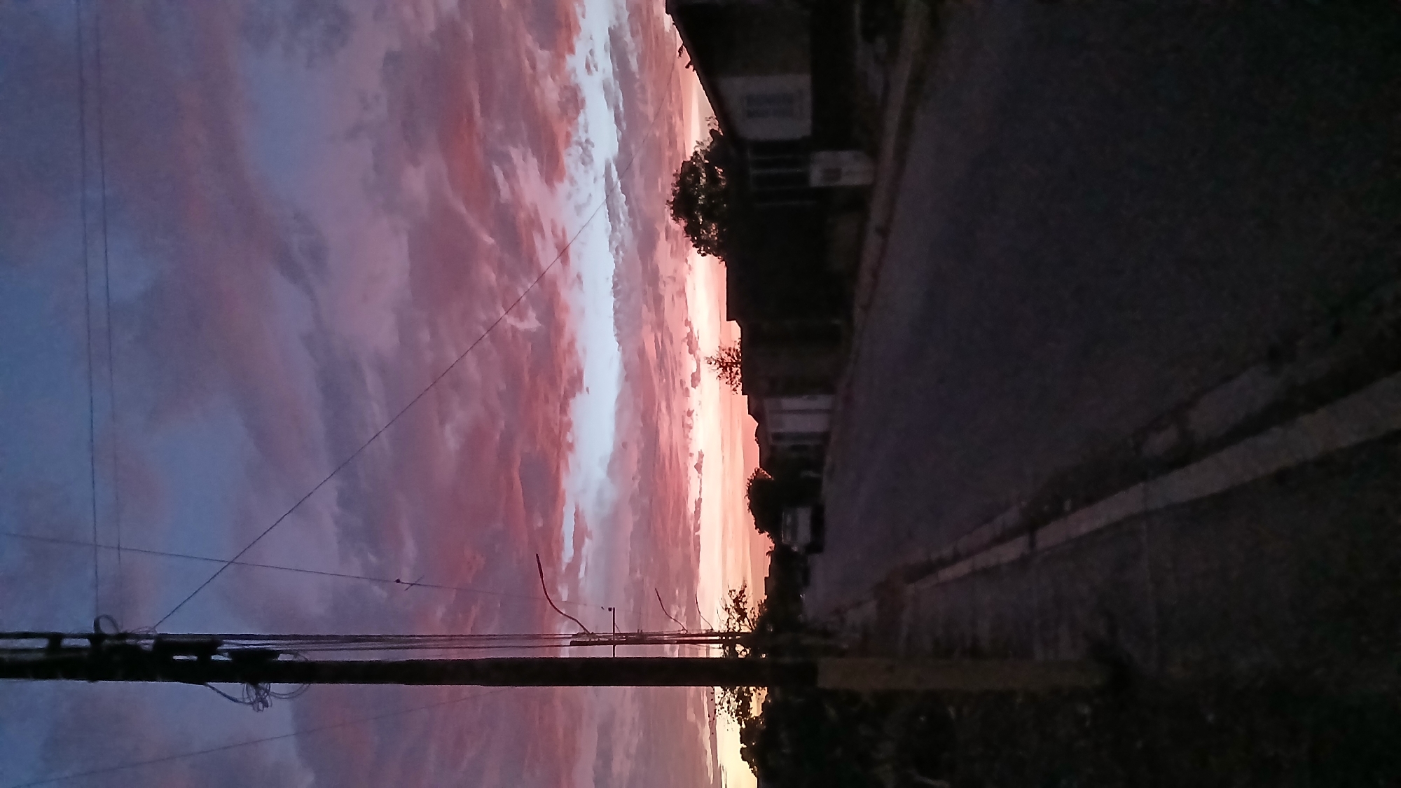 The image shows a street scene in Altagracia, Venezuela, at sunset. The sky is filled with pink and purple clouds. A utility pole with power lines is visible in the foreground on the left. On the right is a road with a curb and houses. The scene is dimly lit.