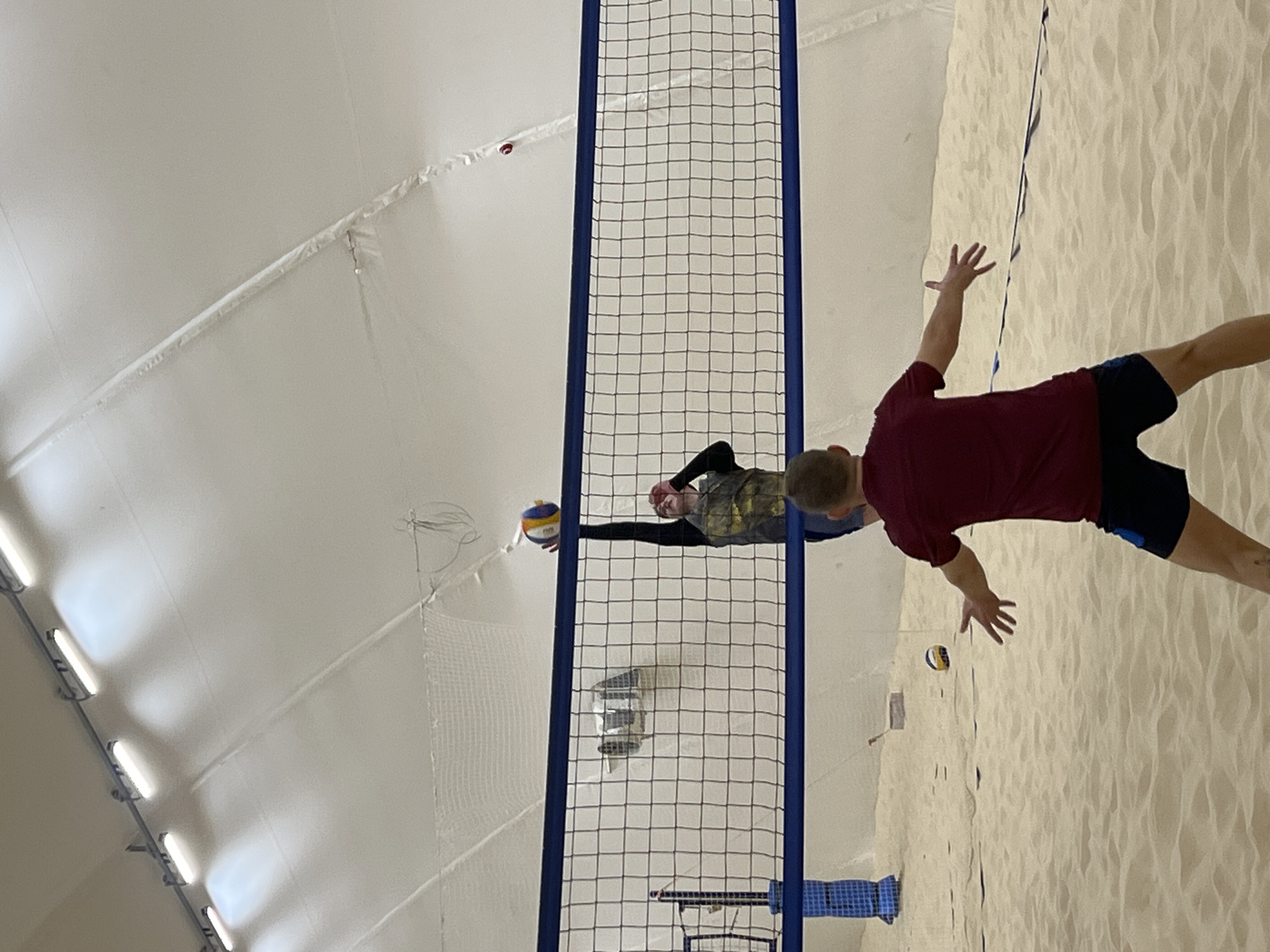 The image shows a volleyball game in progress indoors. Two individuals are positioned on either side of a net. One player in a maroon shirt and dark shorts is attempting to block the ball, while another player on the opposite side is preparing to hit the ball over the net. The playing surface appears to be sand. The indoor facility has a white, tent-like ceiling with lighting fixtures. There are no other distinguishing features that indicate a precise location in Saint Petersburg, Russia.