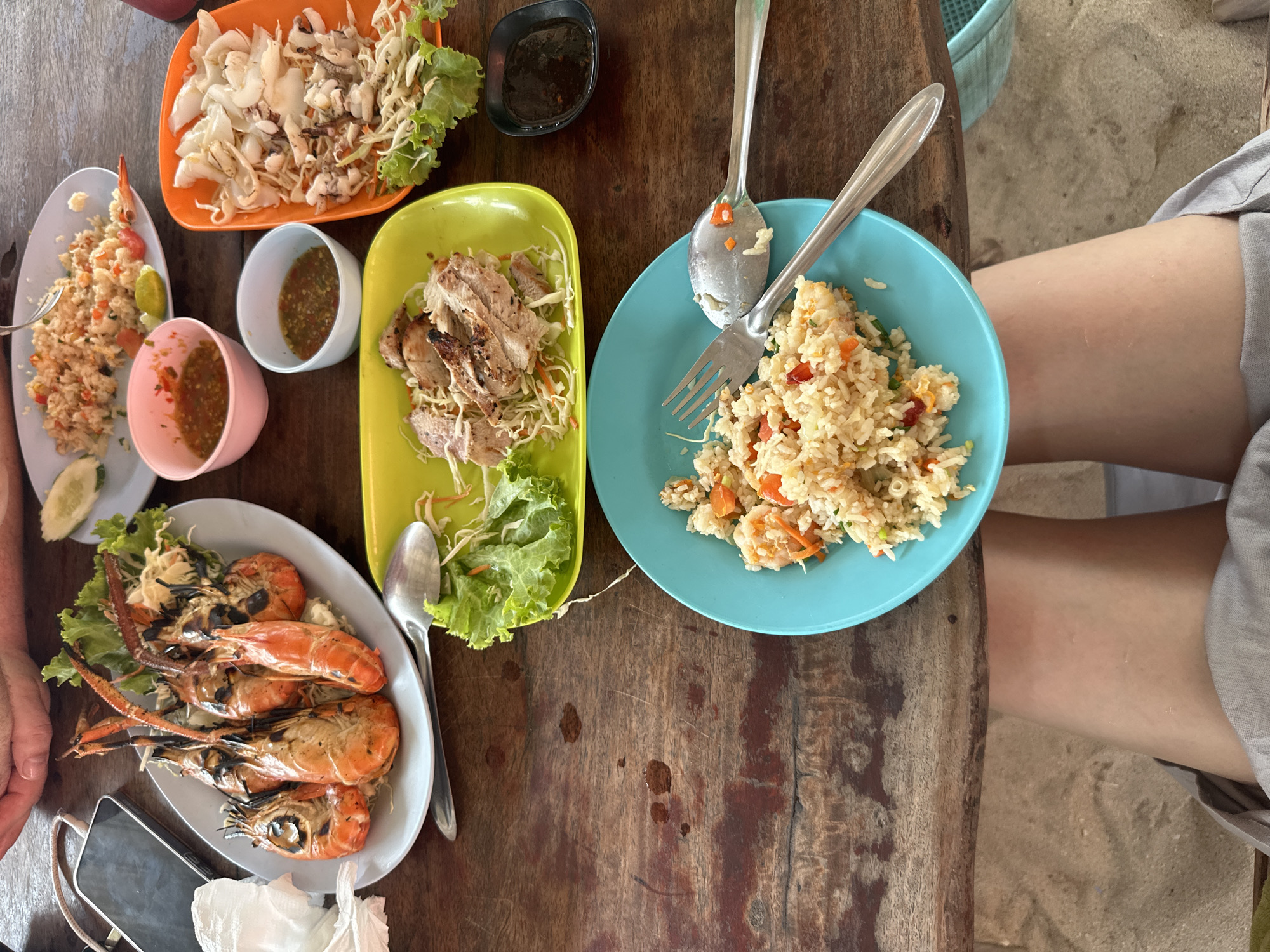 The image depicts a wooden table laden with Thai dishes, possibly taken in Pattaya City, Thailand. There are five plates visible. One plate contains fried rice with shrimp and vegetables. Another plate holds grilled pork with shredded cabbage and lettuce. A third plate displays grilled prawns on a bed of lettuce. A fourth plate has a dish of calamari salad with shredded cabbage and lettuce. There are several small bowls of dipping sauces. Utensils are scattered on the table including a spoon and fork. The table also holds a smartphone, a tissue, and a small purse. A person wearing gray shorts is seated near the edge of the table.