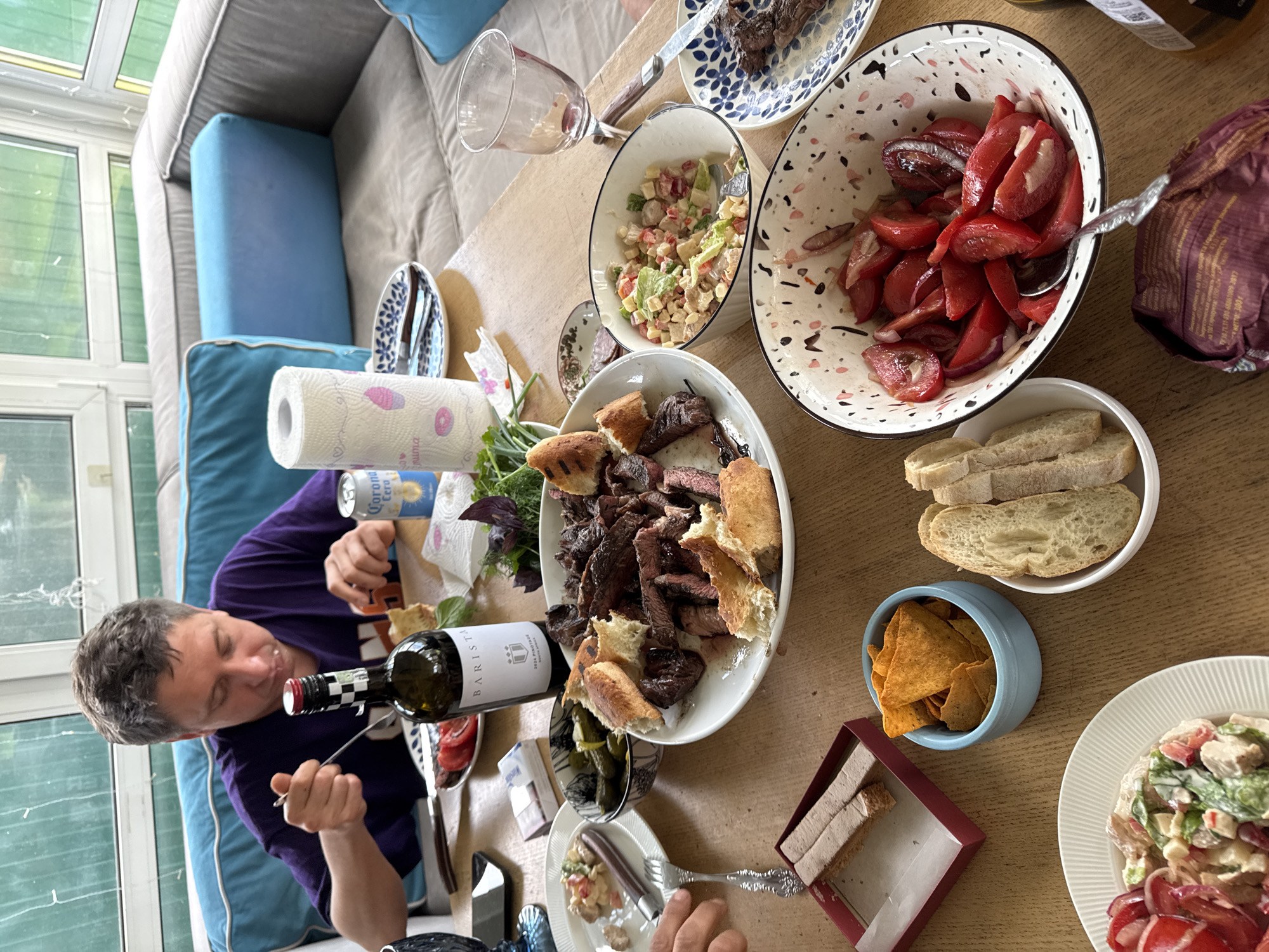 A group of people are gathered around a wooden table, enjoying a meal. The man in the foreground, wearing a purple t-shirt, is eating from a plate of food. Various dishes are spread across the table, including a large platter of grilled meat, bowls of salad, sliced tomatoes, pickles, bread, and a bowl of tortilla chips. A bottle of wine and a can of Corona Extra are also visible. The scene appears to be set in a sunroom or enclosed patio, with large windows looking out onto greenery. The overall atmosphere is one of a casual, convivial gathering. The image was taken in the city of Ptichnoye, Russia.