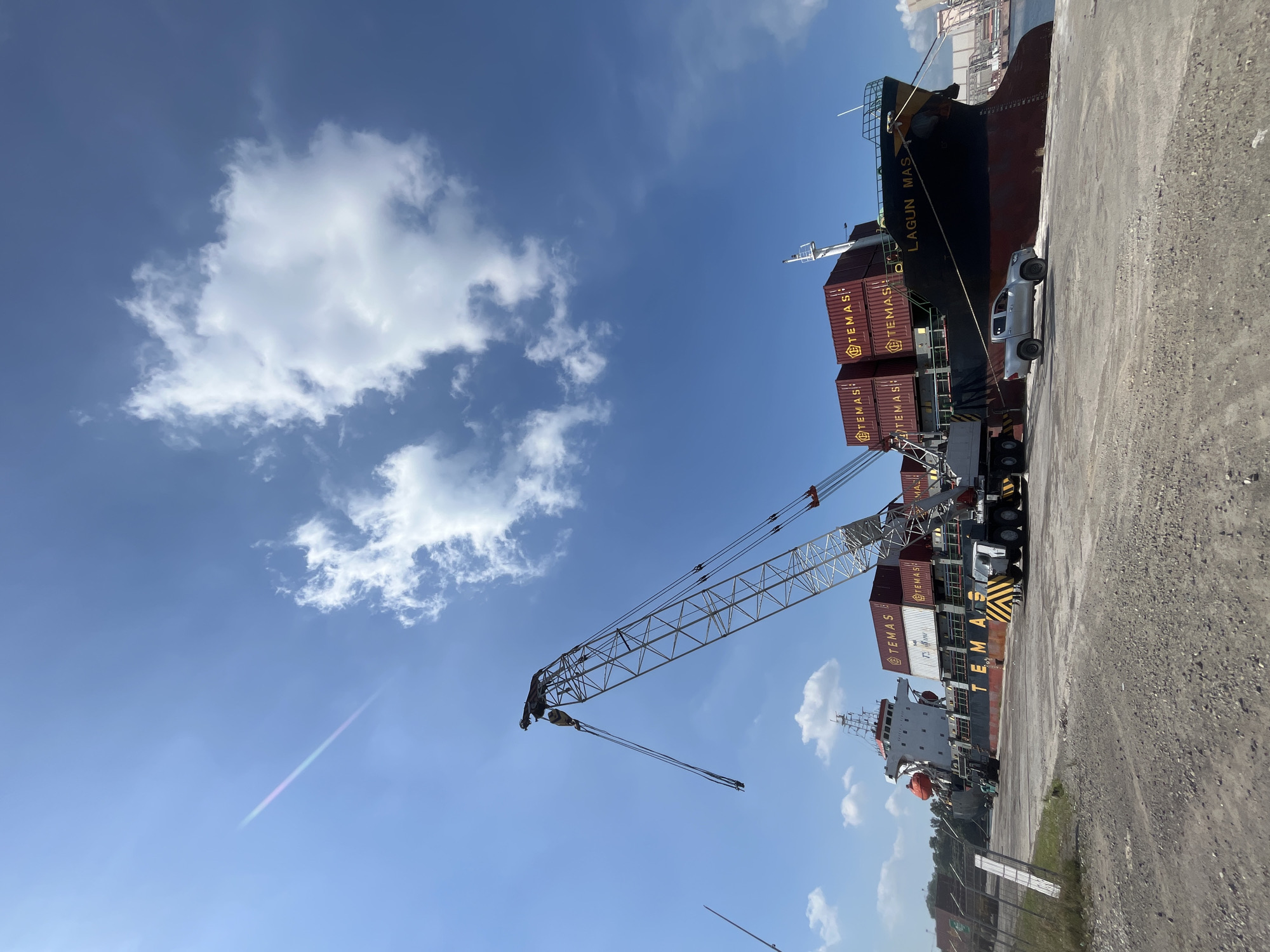 The image shows a docked cargo ship, named "LAGUN MAS," loaded with shipping containers, next to a loading crane at a dockside area in Geulumpangsulu Timu, Indonesia. A silver pickup truck is parked adjacent to the ship. A large crane stands nearby, positioned to move containers. The sky is blue with scattered clouds. The ground is paved.