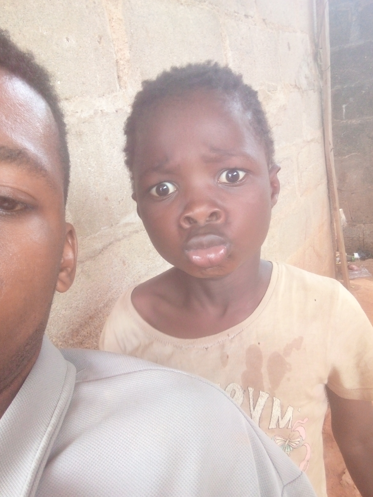 A selfie shows a Black man with short, dark hair and an ear partially visible. He is wearing a light gray, collared shirt. Beside him, a Black child with short, dark hair wears a light yellow t-shirt with a design on the front. The child is making a pouting face. Both subjects are indoors, standing in front of a wall. The location appears to be in an area of Unknown, Unknown.