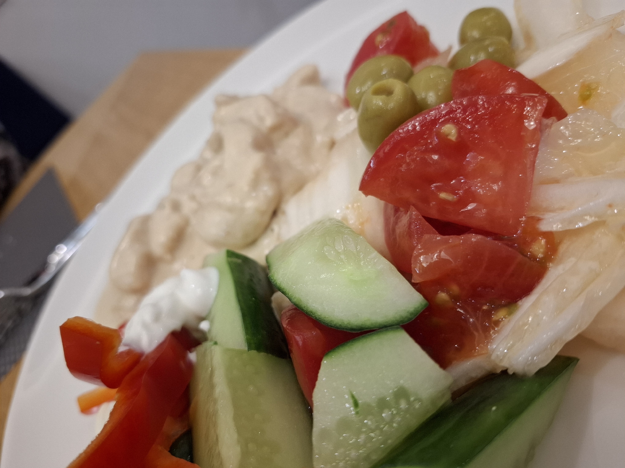 A close-up shot depicts a plate of food. The plate contains a creamy beige sauce, possibly hummus or a similar dip, along with a salad consisting of sliced tomatoes, cucumbers, red bell pepper, green olives, and sliced white onions. A dollop of white sauce, likely yogurt or sour cream, is present near the red pepper. The setting appears to be indoors, on top of a wooden surface. There is insufficient information in the image to confirm the location is Glazov, Russia.