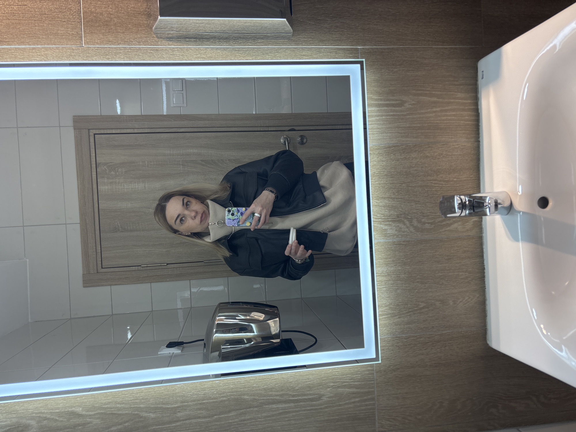 A person is taking a selfie in a bathroom mirror. The bathroom has wood-paneled walls, white tiles, a wooden door, a sink, and a hand dryer. The person is wearing a black jacket and a light-colored hoodie, holding a phone with a patterned case. Based on the bathroom's interior design and the sink model, this is likely a public restroom or a restroom in a commercial establishment located in Russia, Saint Petersburg.
