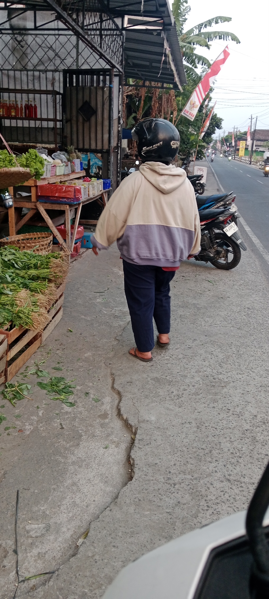 A person wearing a black helmet and a two-toned hoodie is standing on a sidewalk next to a roadside market in Maguwoharjo, Indonesia. The market displays various produce, including leafy greens and pineapples, in wooden crates and plastic containers. The person is facing away from the camera, with their right arm extended slightly. A motorcycle is parked on the street beside the sidewalk. The street is paved and there are other vehicles visible in the distance. Indonesian flags are displayed on poles along the street. The ground is concrete and there are some green leaves scattered on it. The sky appears overcast.