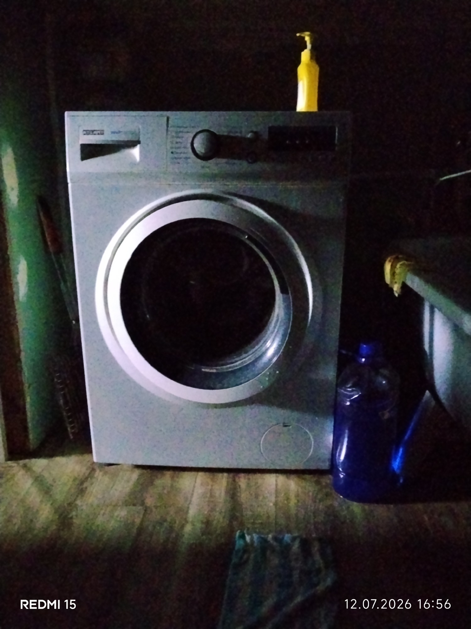 A front-load washing machine is centrally positioned in the frame. It is a light gray appliance with a circular glass door and a control panel on the top left. Above the washing machine, a yellow liquid soap dispenser is visible. To the right of the washing machine, a large blue plastic bottle is placed on a slatted surface, possibly a laundry basket or shelf. The floor appears to be made of wooden planks. The background is dark and indistinct. The lower left corner of the image displays "REDMI 15" and the lower right corner shows "12.07.2026 16:56", indicating the device and date/time the photo was taken. The location is not specified in the image.