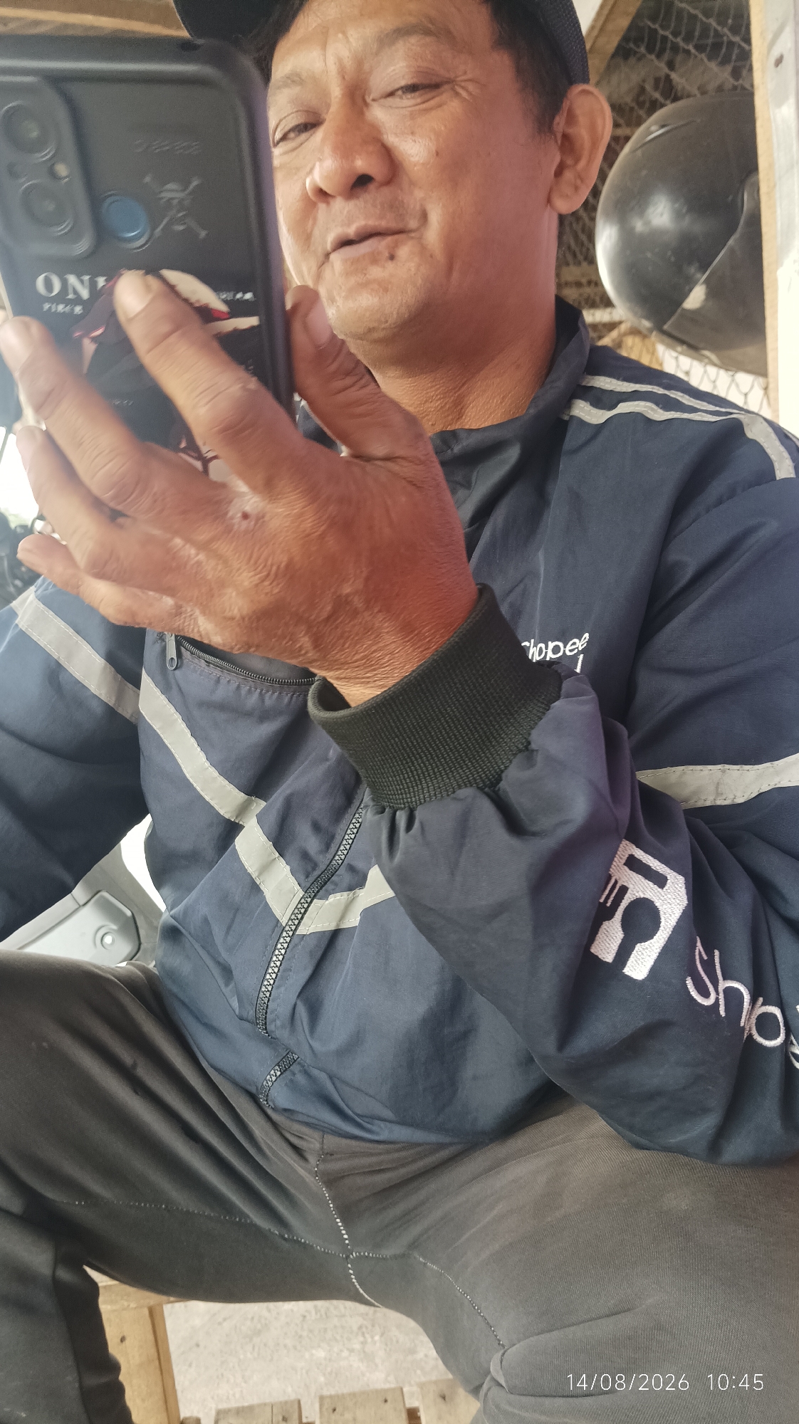 A middle-aged East Asian man wearing a navy blue jacket with grey reflective stripes and a dark baseball cap is holding a black smartphone. He is seated and looking at the phone, with his right hand supporting it. The jacket has "Shopee" and a food icon embroidered on the right sleeve. The background appears to be an outdoor setting, possibly a workshop or storage area, with a black helmet and netting visible. The image also contains a timestamp "14/08/2026 10:45" in the bottom right corner. There is no visual information that specifies the location to be Tasikmalaya, Indonesia.