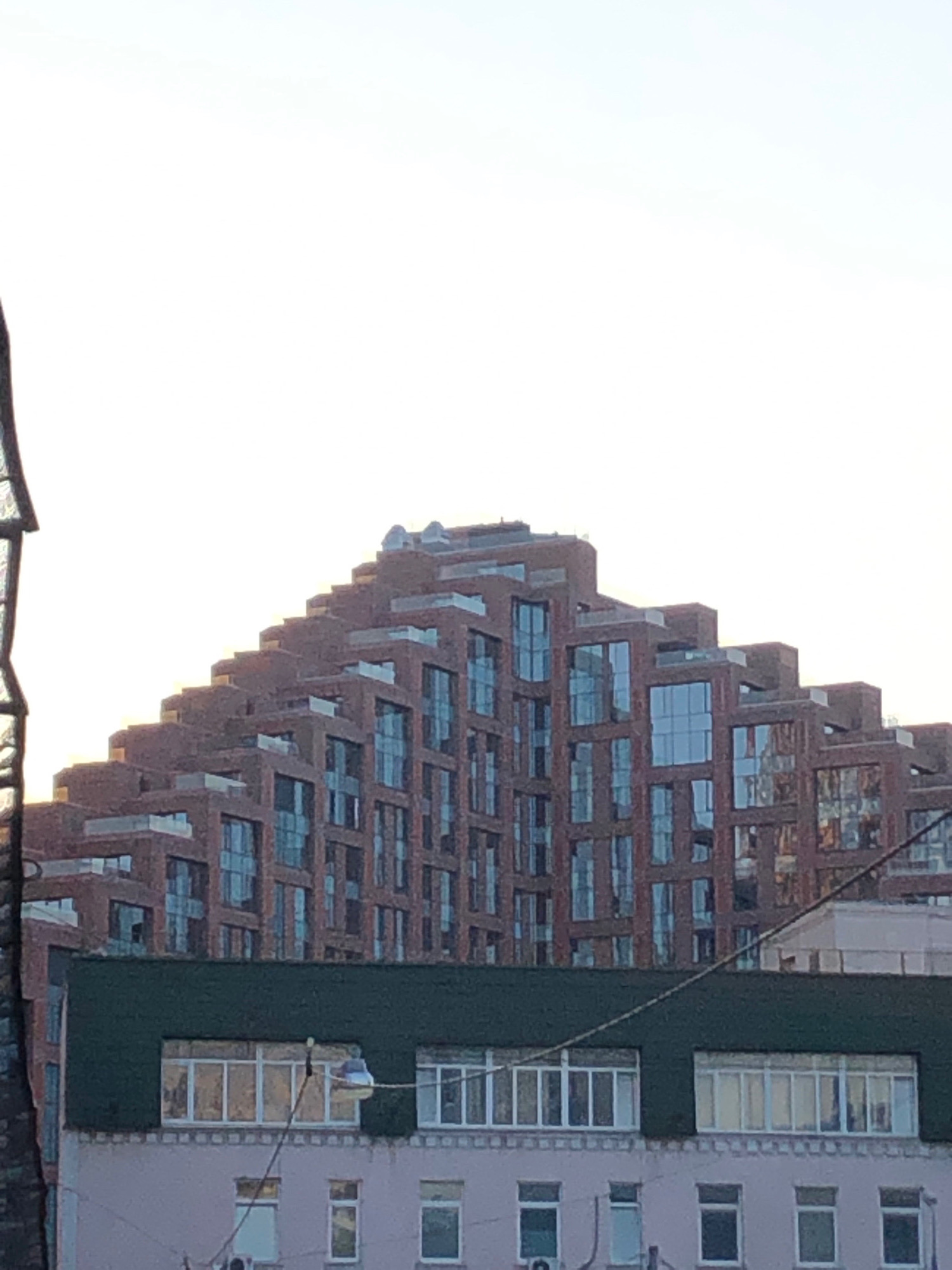 The image captures a view of an unusual, stepped, red brick apartment building with large glass windows, set against a pale sky. In the foreground, a section of a lower, darker green building is visible, also with windows. The overall scene appears to be an urban environment. This building is located in Moscow, Russia.