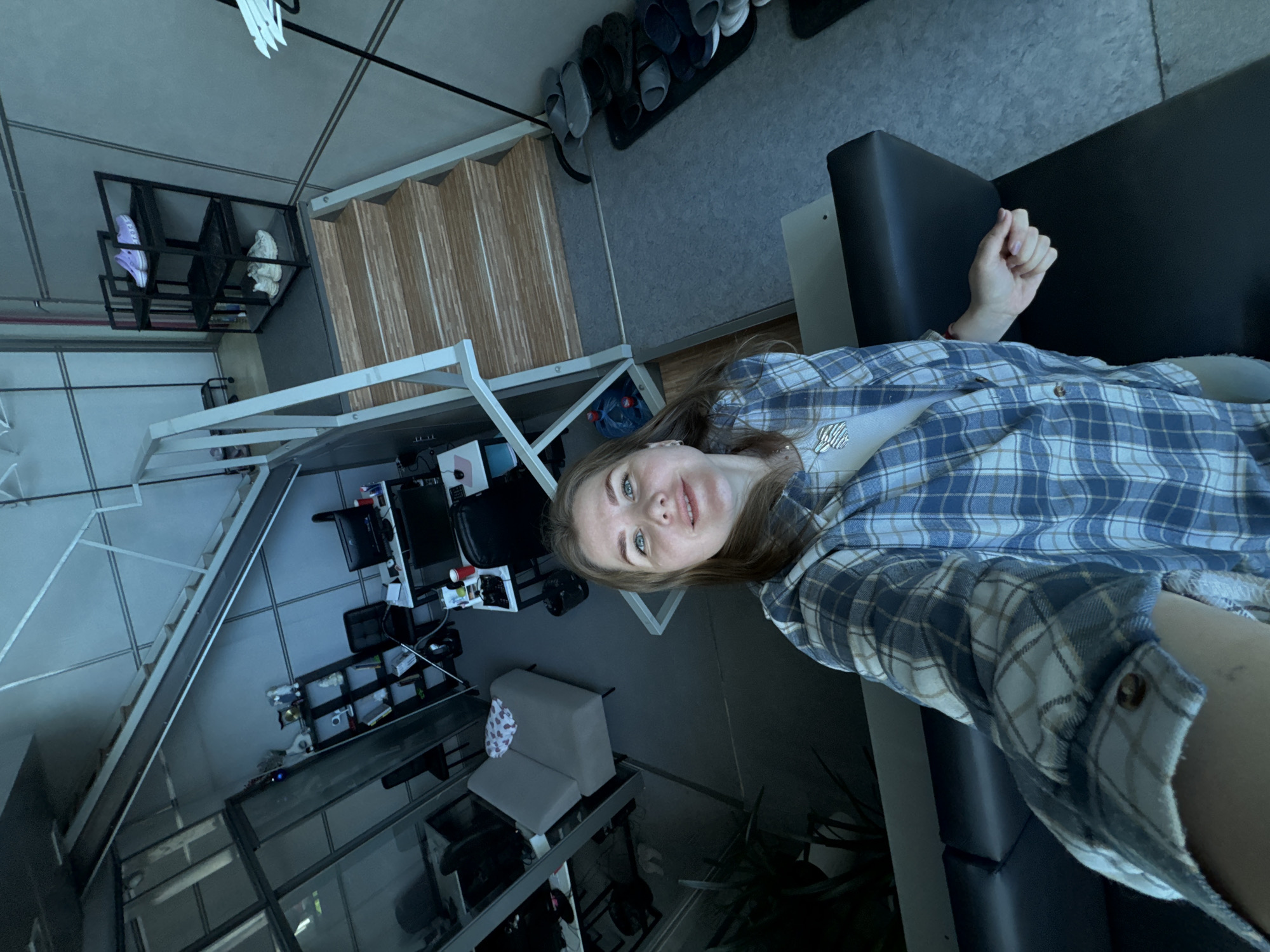 A woman with light skin, light brown hair, and blue eyes is reclining on a black sofa. She is wearing a white undershirt and a blue and white plaid flannel shirt. The photo appears to be a selfie taken indoors, with the camera pointed down at her. The background includes a modern interior with a staircase, office area with computer monitors, shelving, and a row of shoes lined up against a wall. The setting is possibly in Perm, Russia.