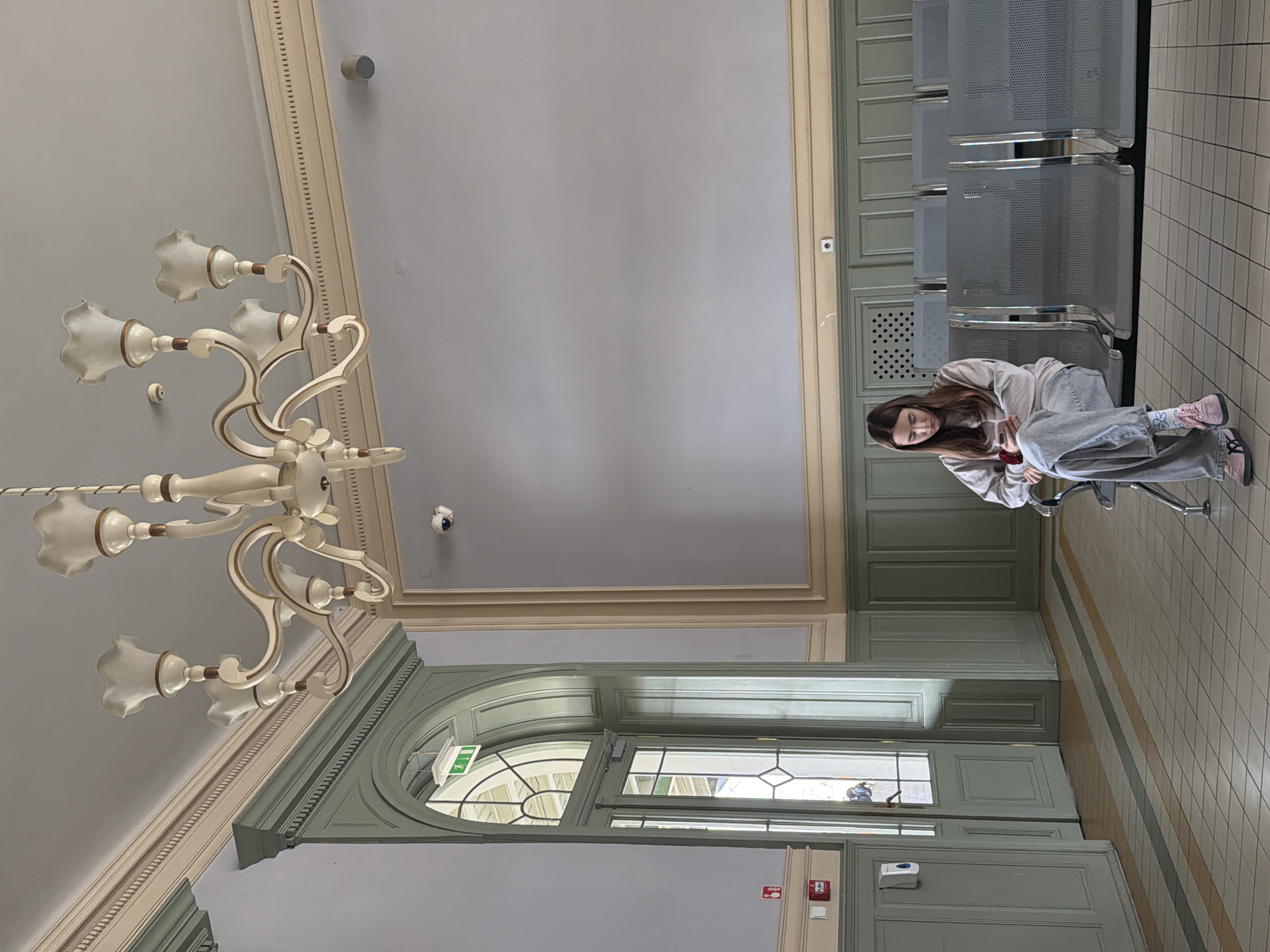A young woman sits in a waiting area in Przemyśl, Poland. She is wearing a light-colored hooded sweatshirt, grey pants, and pink shoes. She is seated on a metal chair with a chrome frame. The waiting area has metal seating aligned against a wall. The wall is painted a light green color. In the background, there is a large window and door structure. A chandelier hangs from the ceiling. The floor is tiled. A security camera is mounted on the ceiling.