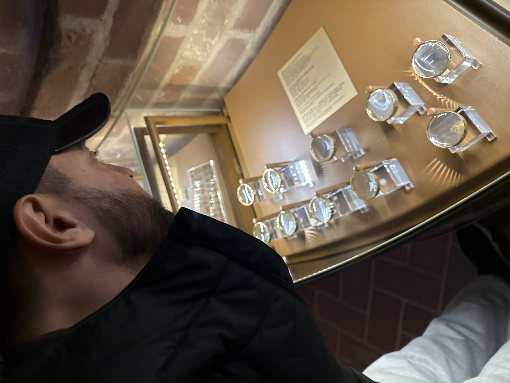 A man wearing a black baseball cap and a black jacket is looking at a display of eyeglasses in a shop. The eyeglasses are arranged on a brown surface, and there is a sheet of paper with text next to them. The shop appears to have brick walls and is possibly located in Moscow, Russia, although this cannot be confirmed from the image. The man is partially visible, focusing on the eyeglasses. The floor is also brick. The lighting is dim and warm. The eyeglasses are of a vintage style with round lenses and unique frame designs.