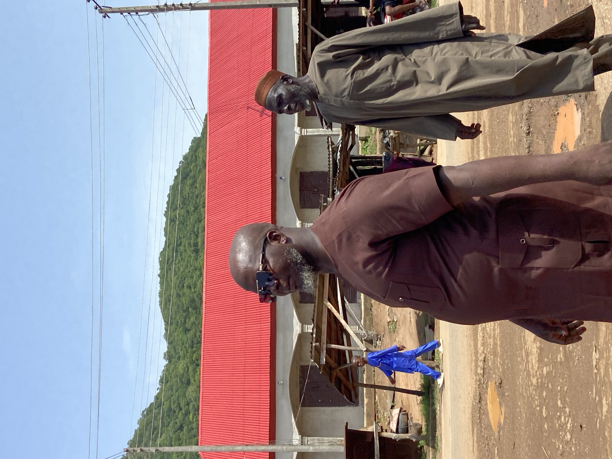 Two men stand facing forward in Mararaba, Nigeria. One man, closer to the camera, wears a brown shirt, sunglasses, and has a beard. He is partially obstructing the view of the other man. The other man wears a brown hat and light brown robe. A red-roofed building is behind them. The sky is blue. Power lines and a forested hillside are visible in the background. A man in a blue outfit and white shoes is visible further in the background. The scene is outdoors. The ground is dirt and gravel.