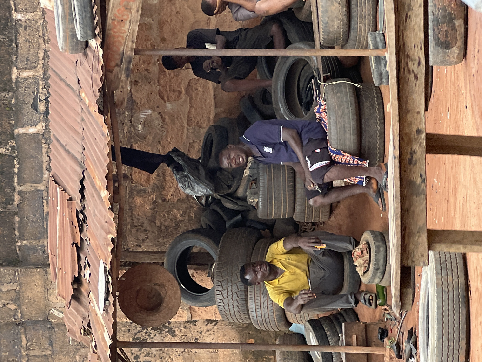 In Nkpor, Nigeria, a tire repair shop operates under a corrugated metal roof, showing signs of rust and damage. Stacks of used tires serve as seating and storage. Three men are present in the scene. The man on the left, wearing a yellow polo shirt and grey trousers, is seated on a tire. He holds a tool in his right hand. A pile of meat is placed on a tire in front of him. In the center, a man in a navy blue polo shirt and black shorts sits on a tire, adorned with a colorful patterned cloth. He appears to be conversing. To the right, a third man, wearing a black t-shirt and shorts, is seated on tires, looking down at his phone. A fourth person, partially visible on the far right, is also seated. The ground is covered in reddish-brown soil, and various tools and discarded items are scattered around the shop. The background consists of a brick wall.