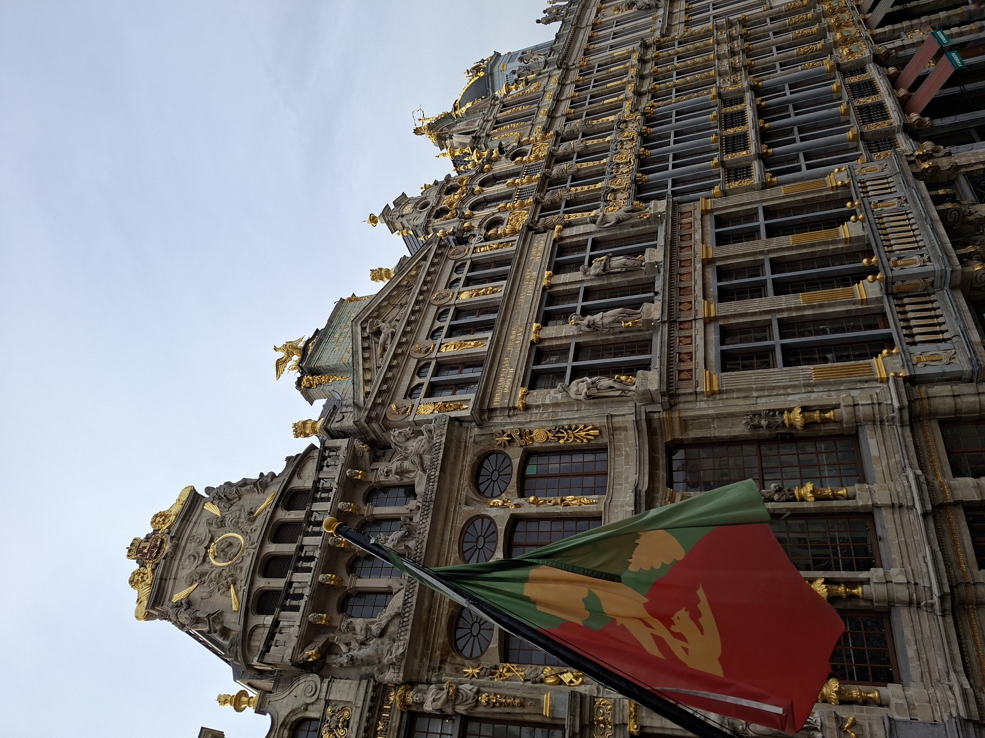 The image depicts the Grand Place in Brussels, Belgium, an iconic square renowned for its ornate architecture. In the foreground, a flag with red, green, and yellow sections is prominently displayed, partially obscuring the view of the buildings behind it. The flag features a yellow crest or emblem against a red background, with a portion of green fabric to its right.
The background is dominated by a row of historical buildings, characterized by their intricate Baroque and Gothic architectural styles. These structures are adorned with extensive gold detailing, including sculptures, decorative elements, and gilded statues atop their gables and facades. The buildings exhibit multiple stories with numerous windows, many of which are dark and reflect the overcast sky. Some of the architectural features include prominent cornices, balconies with decorative railings, and classical sculptures in niches. The sky above is a pale, uniform gray, suggesting a cloudy day. The overall scene conveys a sense of historical grandeur and architectural splendor.