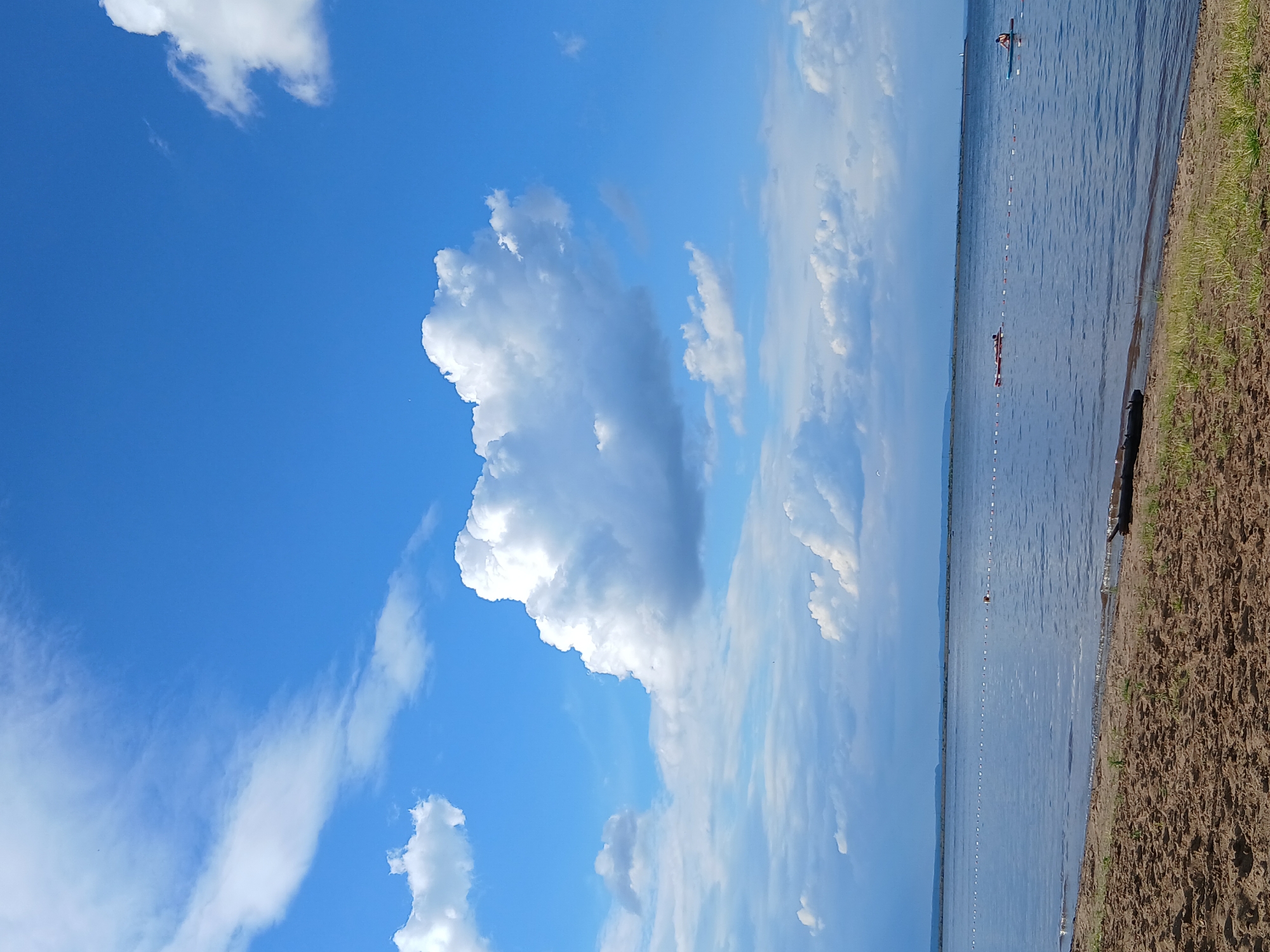 The media file depicts a scenic outdoor scene with a focus on a body of water, a shoreline, and a bright blue sky filled with clouds. The perspective suggests a slightly elevated viewpoint looking down towards the water.
The subjects in the scene include two individuals on the water. One person is in a kayak, and another appears to be on a paddleboard. They are positioned in the distance, near a line of buoys that mark a boundary in the water. The water itself is calm with gentle ripples.
The shoreline on the right side of the frame is composed of sandy or earthy ground with sparse, dry grass. The sky is a dominant feature, with large, fluffy white clouds against a vibrant blue background. The lighting suggests it is daytime, likely mid-morning or afternoon, with the sun illuminating the clouds and water. The overall atmosphere is peaceful and recreational, suggesting a summer day activity.
The location is identified as Chaikovsky, Russia. There is no visible text within the media file. The scene evokes a sense of tranquility and outdoor enjoyment.