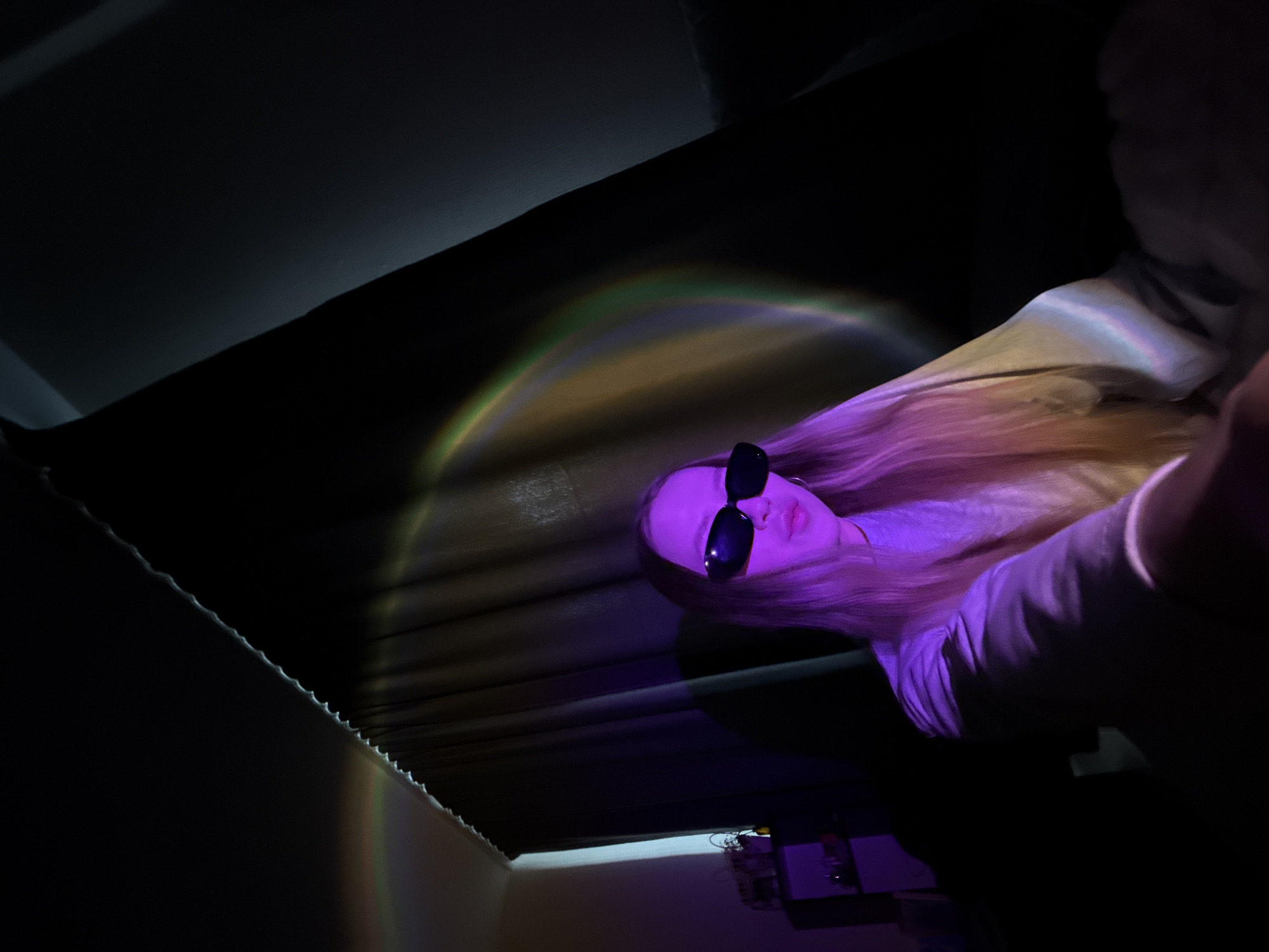 This indoor image, taken from a low angle, features a person with long blonde hair and sunglasses positioned in the upper right. They are illuminated primarily by a striking purple light, casting a vibrant hue on their face, hair, and light-colored top. A prominent rainbow-like light projection arcs across a dark, possibly curtained or upholstered background, extending partly onto the person's hair, showcasing colors ranging from green to yellow and orange.
The scene appears to be set at night or in a dimly lit room, with artificial light sources creating a moody and stylized atmosphere. In the lower right corner, several electronic devices or objects are faintly visible on a surface, also bathed in the purple light. The textured edge of what appears to be a curtain or similar fabric runs diagonally across the left side of the frame. The person is posing for the camera, suggesting a casual portrait or selfie in an ambiently lit setting. No visible text is present. The location context for this image is Chaikovsky, Russia.