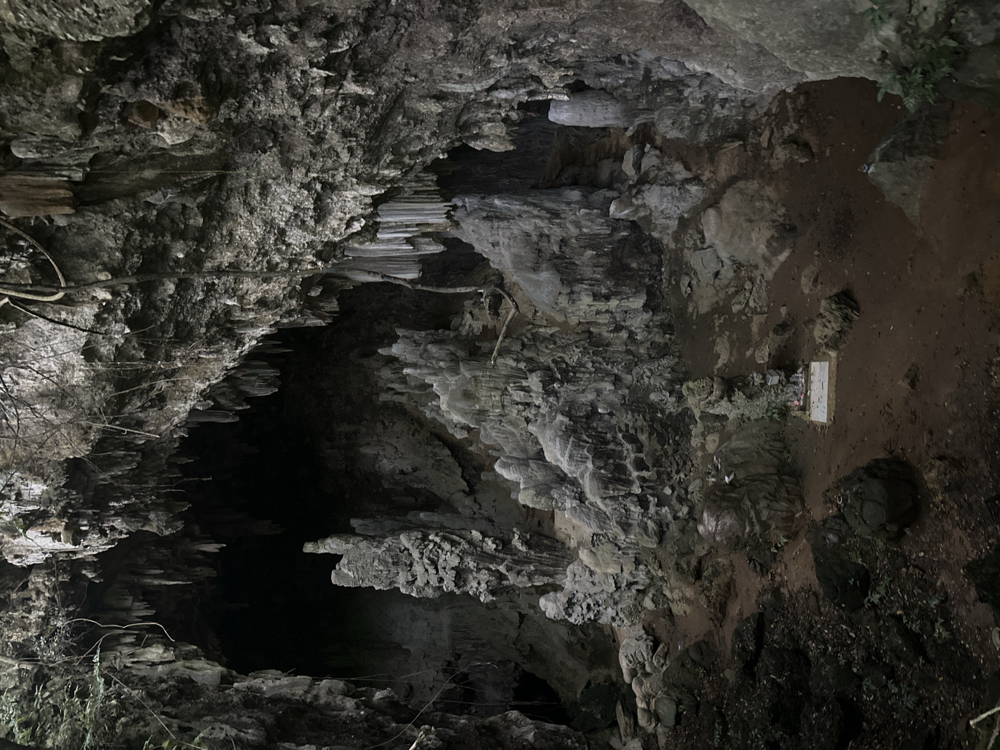 The image depicts the interior of a cave, likely in or near Xóm Khán, Vietnam, characterized by its natural rock formations and dim lighting. Stalactites hang from the cave ceiling, and stalagmites rise from the floor, creating a textured, almost sculptural landscape. The ground is composed of dark soil and scattered rocks, with some patches of sparse vegetation. In the mid-ground, a small, rectangular stone altar with offerings is visible, suggesting this location may be a place of worship or cultural significance. The cave entrance or a deeper section of the cave is visible in the background, appearing as a dark, cavernous opening. There are no people visible in the image.