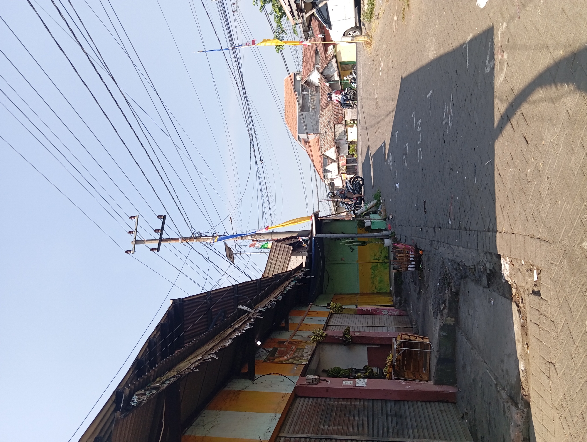 The image depicts a street scene in Kepulungan, Indonesia, under a clear sky. On the left, a row of shops with corrugated metal roofs and roll-up doors line the street. One shop is painted with vertical stripes of yellow, orange, and light blue, with a partially visible sign that reads "DEWI...". Bananas are displayed for sale near the entrance of this shop, hanging from the awning and placed on a wooden crate.
The street itself is paved with interlocking brick pavers. There are markings painted in white on the pavement, some of which are discernible as numbers and letters, such as "17", "27", and "10".
Several individuals are present. Two people are riding a motorcycle in the middle of the street, heading away from the camera. Another person is on a motorcycle parked on the left side of the street. A third person is seated on a motorcycle further down the street on the right.
Power lines crisscross overhead, supported by wooden utility poles. Flags, including a yellow one and one with Indonesian national colors, are visible on poles along the street. Buildings with terracotta-tiled roofs are visible in the background, suggesting a residential area behind the shops. A white car is parked on the right side of the street. The overall impression is of a typical street in a developing urban or semi-urban area.