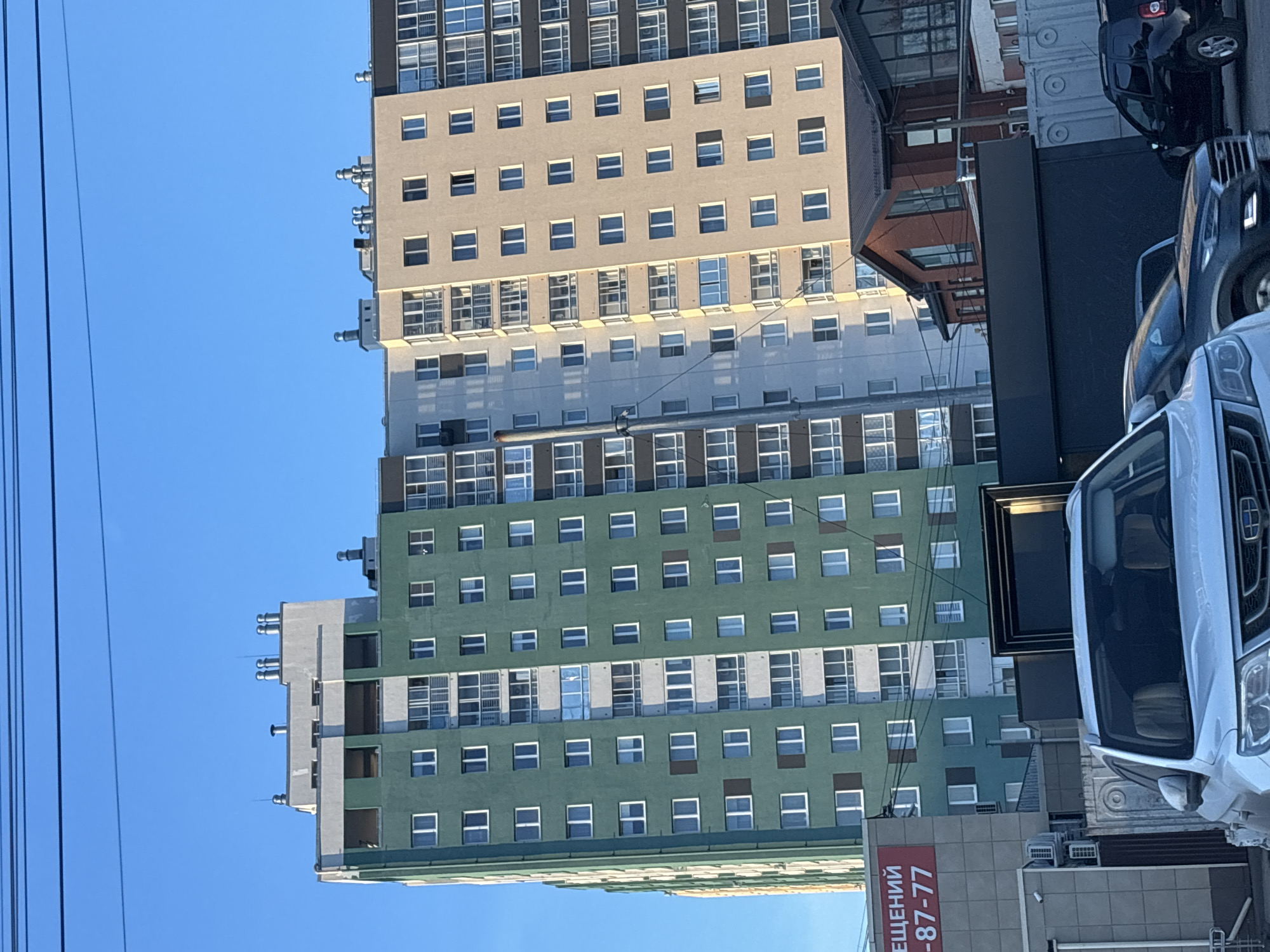 The image depicts a vibrant urban scene dominated by a tall, multi-story building under a clear blue sky. The building, likely residential or mixed-use, features distinct sections painted in different colors: the upper floors are a light brown/beige, followed by a grey/white middle section, and the lower floors are a striking green. Each section is characterized by a grid of numerous square windows, with some floors having larger, recessed window designs. Ventilation or HVAC units are visible on the rooftops and ledges of the building's different sections.
To the right of the main building, a street or parking area is visible with several vehicles. A white van or SUV occupies a prominent position in the immediate foreground on the right, with a dark-colored SUV partially visible behind it. Other, lower-rise buildings, possibly commercial or older residential, can be seen in the background on the right.
Overhead power lines or utility wires stretch across the bright blue sky on the left side of the frame. The clear sky indicates a sunny day.
A notable detail is a red sign with white Russian text located at the bottom right, below the green section of the tall building. The visible text reads "ЩЕНИЙ -87-77", which is likely part of a phrase like "ПОМЕЩЕНИЙ" (meaning "premises" or "spaces") followed by part of a phone number, suggesting an advertisement for property.
The setting is an urban environment, likely in Izhevsk, Russia, characterized by modern architecture and street activity.