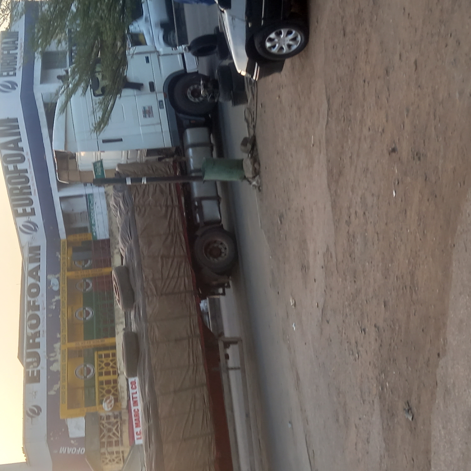 The image shows a street scene in Dadi Riba, Nigeria. A Eurofoam building is visible, alongside a truck loaded with covered cargo. A parked black car is also present. The road appears to be unpaved. No people are clearly visible in the image.