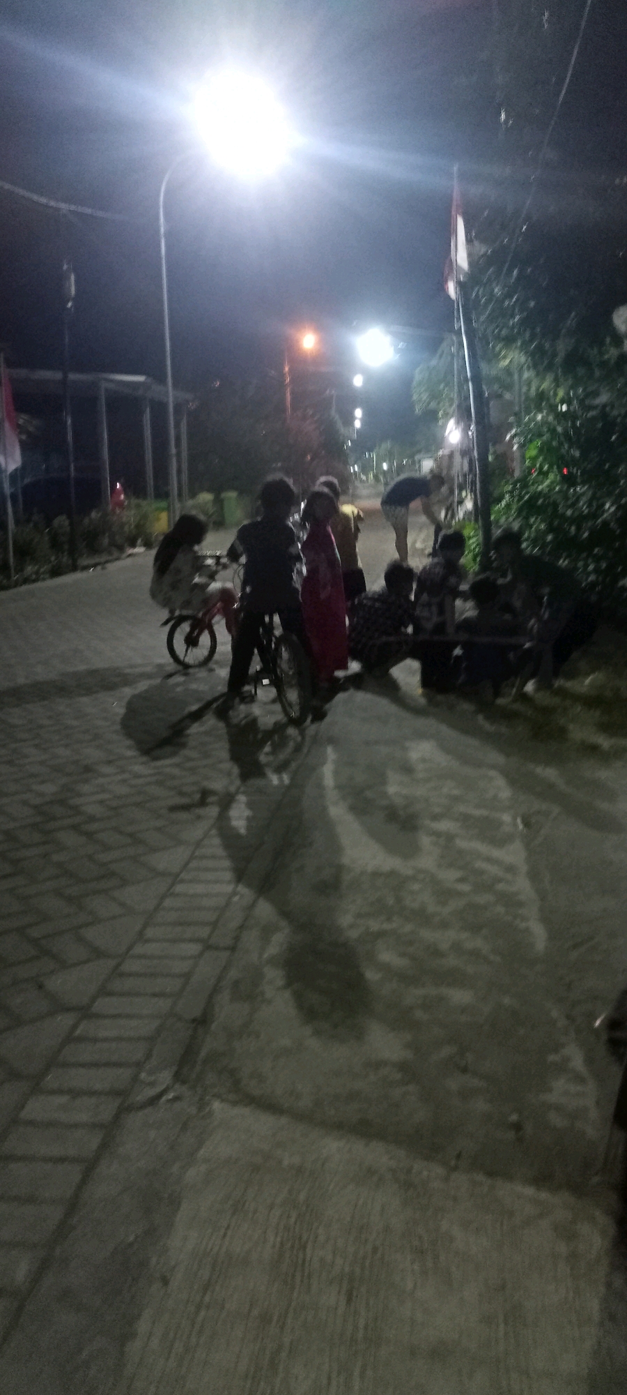 In Gempolklutuk, Indonesia, a group of people, predominantly children, are gathered outdoors at night, illuminated by streetlights. The scene takes place on a paved walkway with a textured pattern and a concrete area adjacent to it. Several individuals are on or near bicycles. In the foreground left, a child rides a small red bicycle. Next to them, another child stands by a black bicycle. Further back, a cluster of figures are interacting near a bench. One person is bent over what appears to be a bicycle, possibly making repairs or adjustments. Another individual stands beside them. A flag, seemingly the Indonesian flag due to its red and white colors, is visible on a pole in the background, as are trees and some buildings with covered structures. The lighting from the streetlights casts long shadows on the ground.