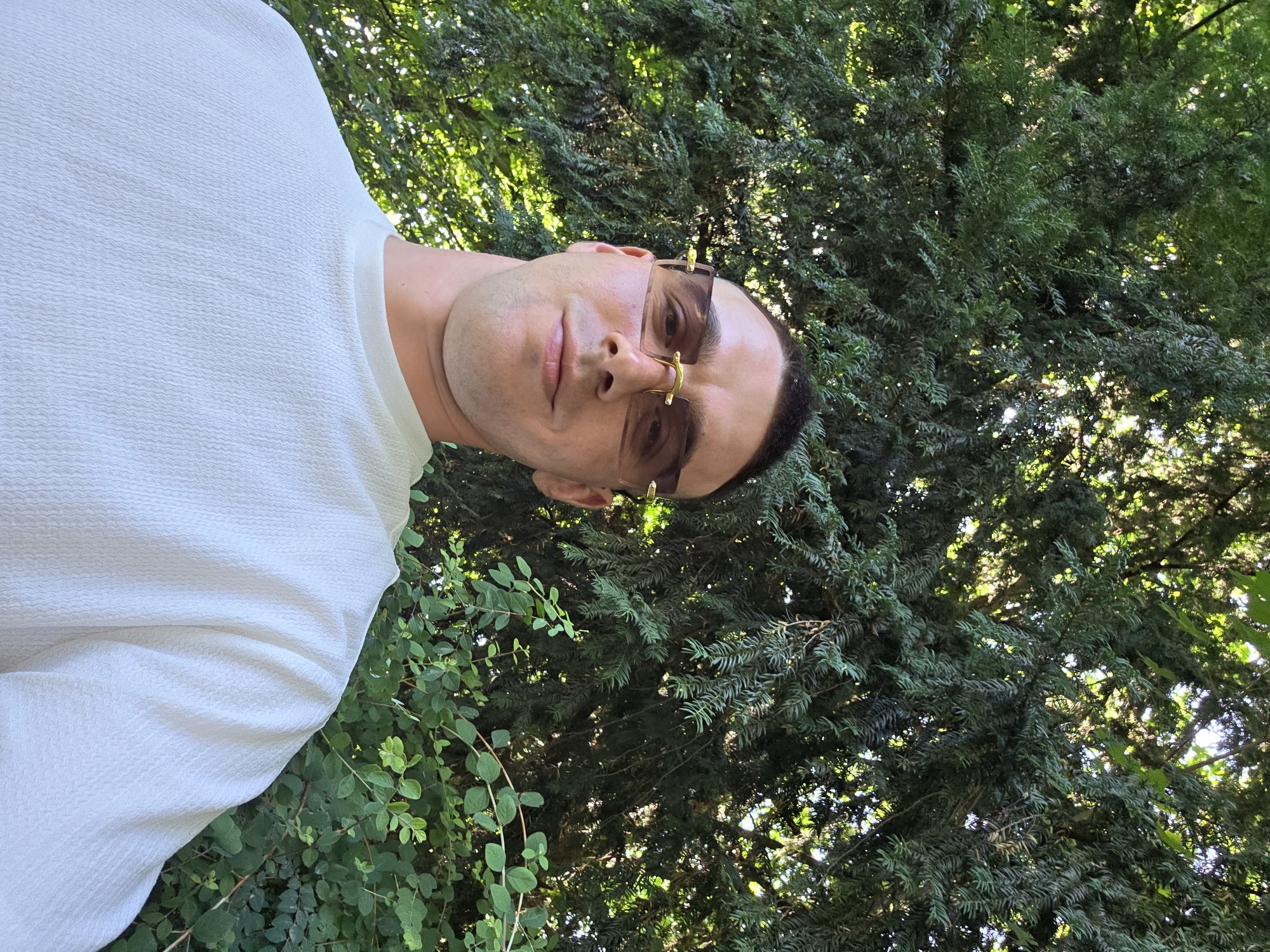 A male individual is depicted in a close-up, frontal portrait, positioned in the lower center of the frame against a natural, leafy background. He appears to be in his late 20s to early 30s, with short, dark hair and a light tan complexion. He is wearing rimless, rectangular sunglasses with brown-tinted lenses and gold-colored metal accents on the bridge and temples. His expression is a slight smile. The individual wears a light-colored, possibly white or cream, crew-neck t-shirt with a distinct textured, wavy pattern on the fabric. The background is dominated by dense green foliage, consistent with large trees and smaller bushes, creating a dappled light effect with sunlight filtering through the leaves. The setting is outdoors, likely a park or garden. The location is in the city of Unknown, Unknown.