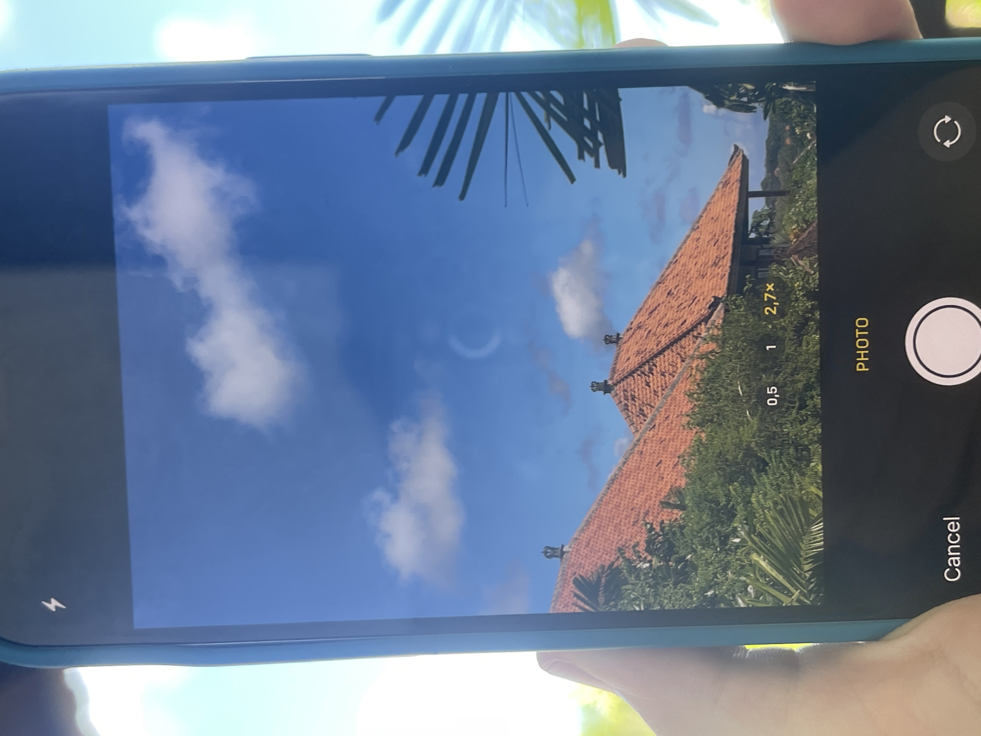 A smartphone held in someone's hand displays the camera app's interface, capturing a scene of a red-tiled roof with greenery and a blue sky with scattered clouds. The phone screen also shows the camera app's controls, including zoom levels, the photo button, and the cancel button. The phone case is light blue. The location is unknown.