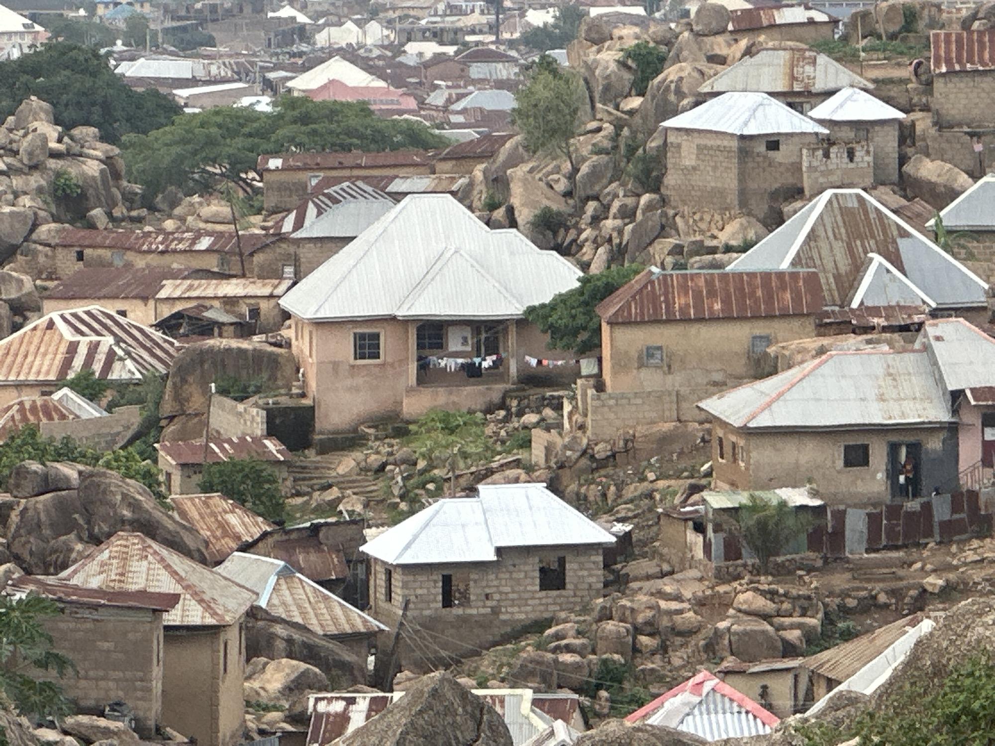 This image captures a dense residential settlement built into and around a rocky, hilly landscape, characteristic of Jos, Nigeria. Numerous single-story homes, predominantly constructed from concrete blocks or rendered earth in shades of brown and beige, are scattered across the terrain.
The roofs are a mix of corrugated metal sheets, with some appearing rusted in reddish-brown tones and others gleaming silver, indicating newer construction or repairs. Large, natural rock formations are an integral part of the environment, with houses nestled between, on top of, and alongside these imposing boulders, demonstrating how the community has adapted to the challenging geology.
Sparse green vegetation and trees are visible, breaking up the earthy tones of the rocks and buildings. In the mid-ground, laundry hangs to dry on a line outside a central house with a bright silver roof, suggesting ongoing daily domestic activities. Further to the right, a figure, likely a child, can be seen in the doorway of another house. Various makeshift fences and partitions made of corrugated metal are also discernible throughout the scene.
The image portrays a daytime setting, under what appears to be an overcast or hazy sky, providing soft, diffused lighting with no strong shadows. There is no discernible text visible in the image. The scene conveys a sense of a vibrant, lived-in community ingeniously utilizing its natural surroundings.