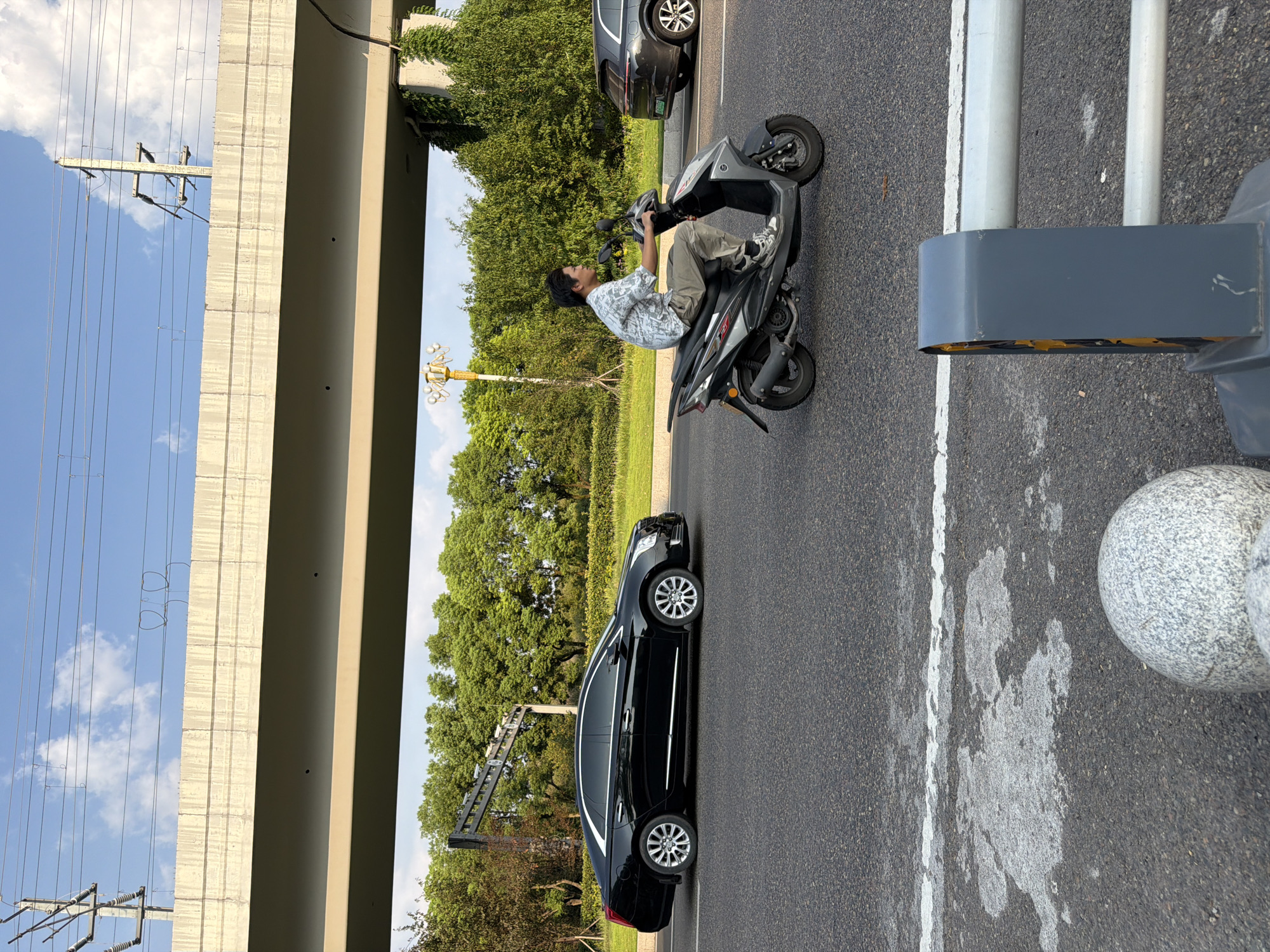 A man rides a grey scooter on an asphalt road. He is wearing a light-colored patterned shirt and tan pants. Behind him, a black Cadillac sedan drives in the same direction. In the distance, a dark-colored SUV is parked. The road is bordered by a grassy median and lush green trees. An elevated concrete overpass with overhead power lines spans the scene. The sky is blue with scattered white clouds. The image was taken in Jiangjin, China.