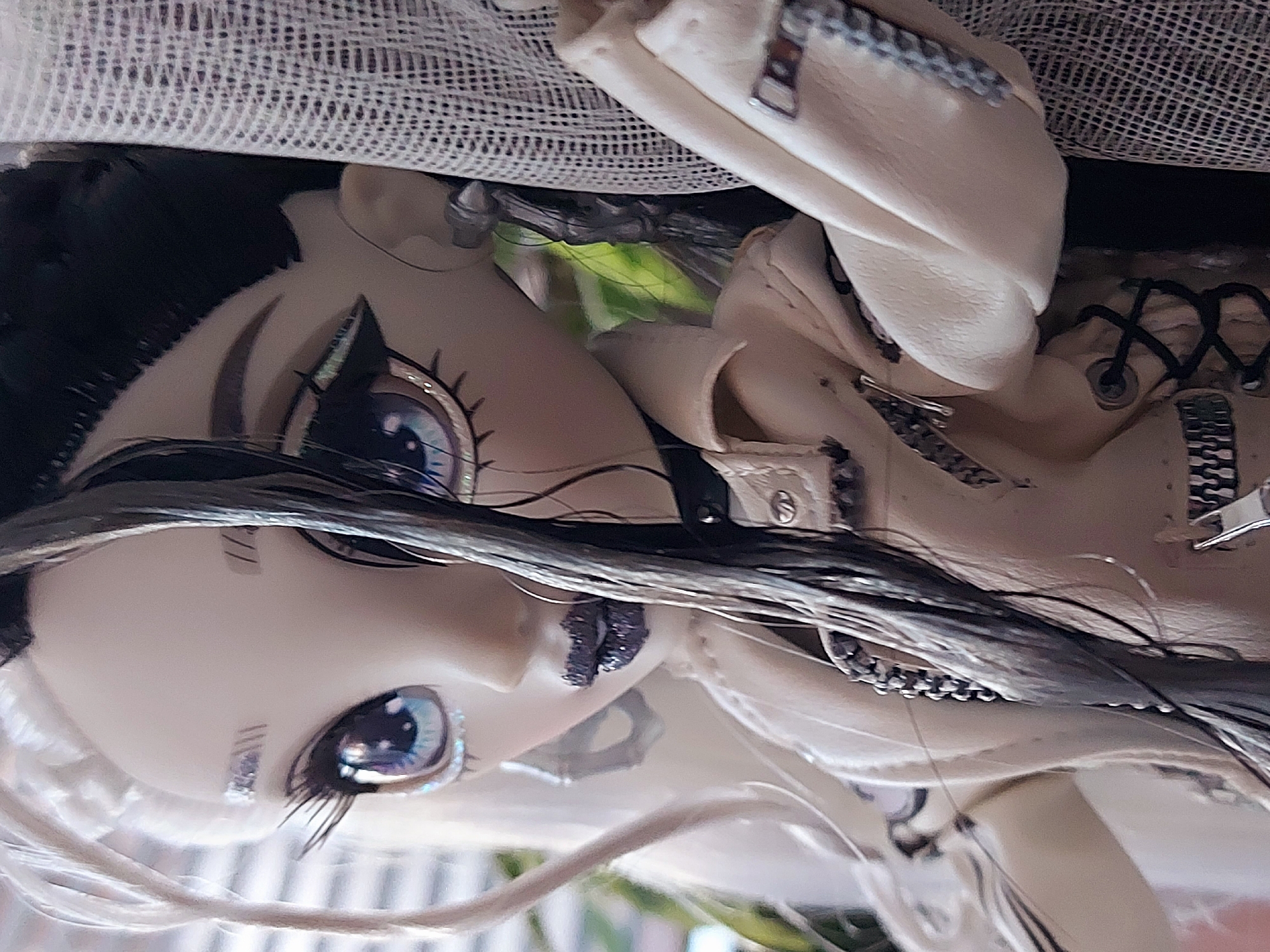 The image is a close-up, three-quarter portrait of a fashion doll set in an indoor environment in the city of Chaikovsky, Russia. The doll features a pale complexion with distinct split-dyed hair: white-blonde on the viewer's left and dark gray-black on the viewer's right, with some strands falling across its face. Its large, light blue eyes exhibit heavy black winged eyeliner, long eyelashes, and sparkling tear duct details. The lips are covered in glittery dark purple or black lipstick. Stylized double-line marks are visible on one eyebrow, and a silver stud along with a geometric dangle earring adorn the visible ear. The doll is dressed in a light beige faux leather jacket with multiple silver zipper accents, chain embellishments, and black lacing. It is positioned adjacent to a panel of light, open-weave mesh fabric on the right. The blurred background reveals vertical window blinds and hints of green foliage outdoors.